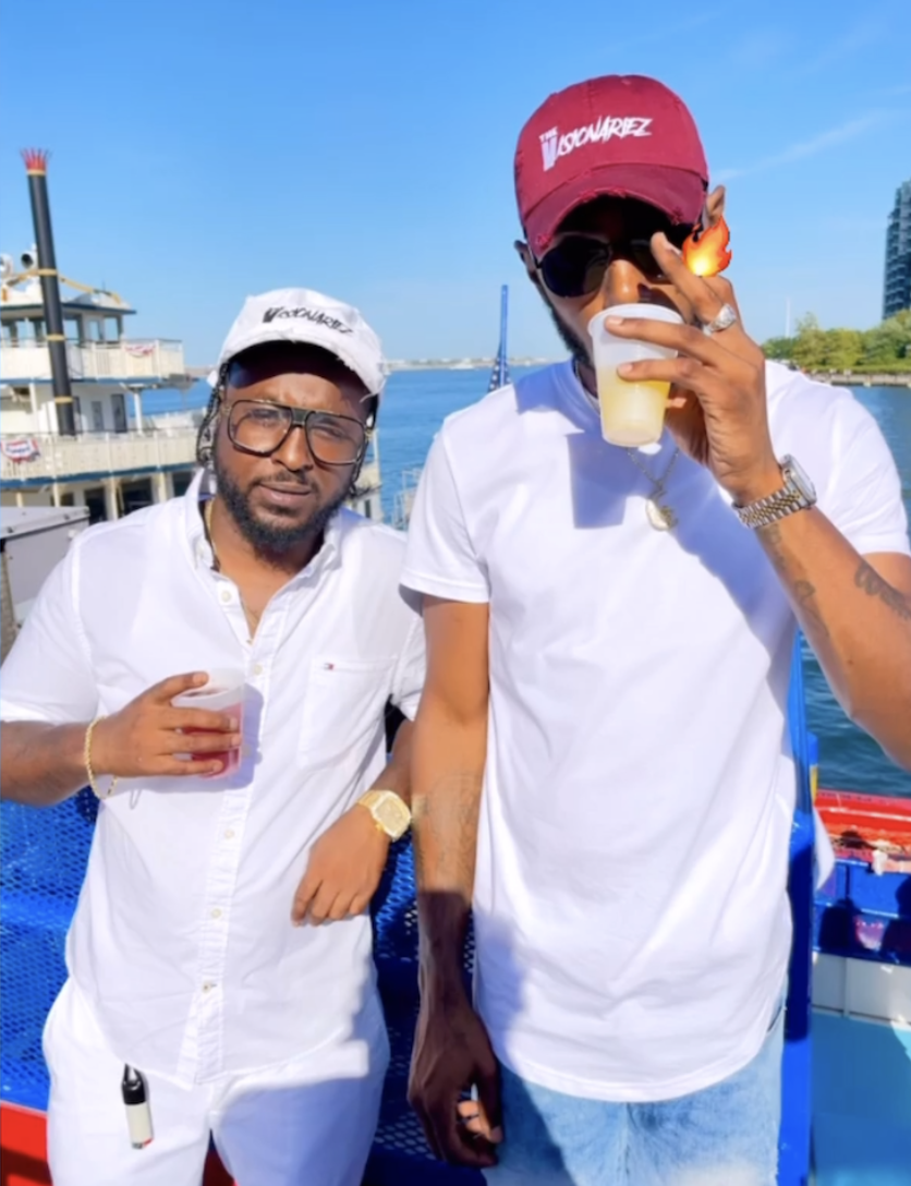 Two men standing by a waterfront holding drinks, wearing casual white shirts and caps, with blue sky and water in the background.The Visionariez