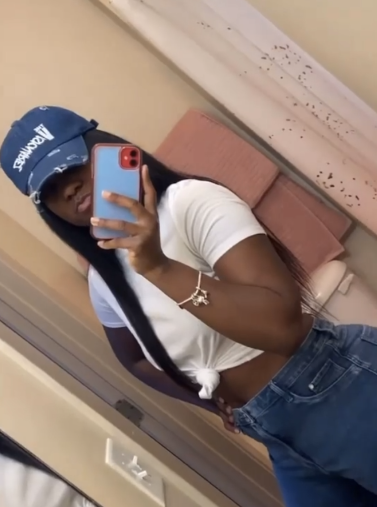 A woman taking a mirror selfie with her phone, wearing a blue cap The Visionariez, white t-shirt tied at the waist, and blue jeans.