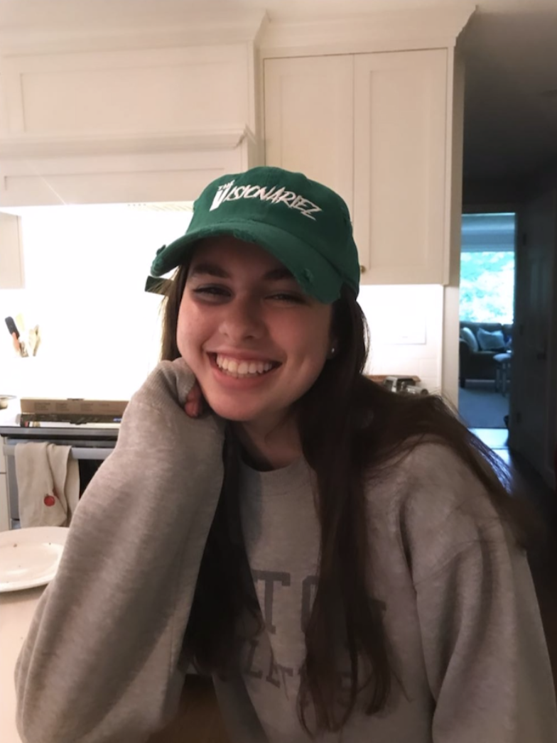 A young woman with long brown hair, smiling and wearing a green cap with white text, sitting in a kitchen. The Visionariez'