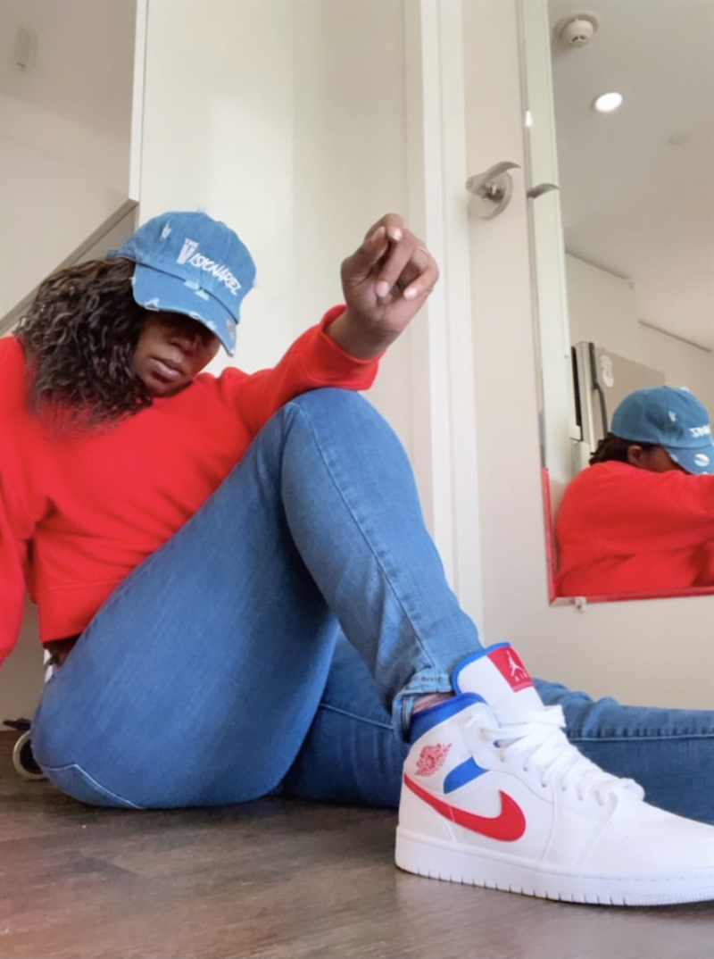 A person sitting on the floor wearing a red sweatshirt, blue jeans, and white sneakers with red and blue accents, in front of a mirror, with their head tilted down and a blue cap on, in a room with white walls and a wooden floor. The Visionariez