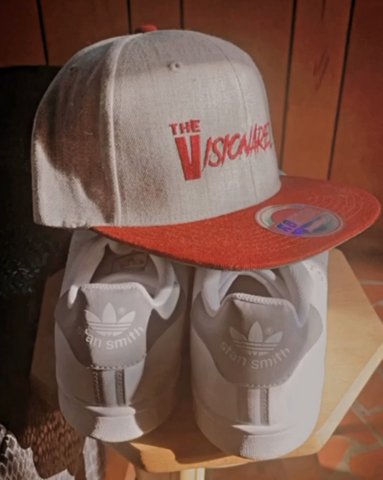 A beige and orange snapback hat with the text 'The Visionariez' in red, a pair of white Adidas Stan Smith sneakers, and a wooden surface on which they are placed.
