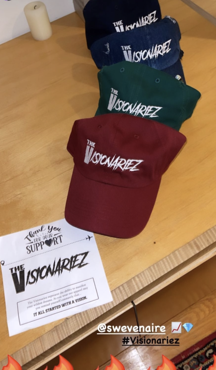 Four baseball caps in black, navy, green, and red, all with the words 'The Visionariez' embroidered on the front, are displayed on a wooden table. A printed note with similar branding and a handwritten message with social media tags is also on the ta