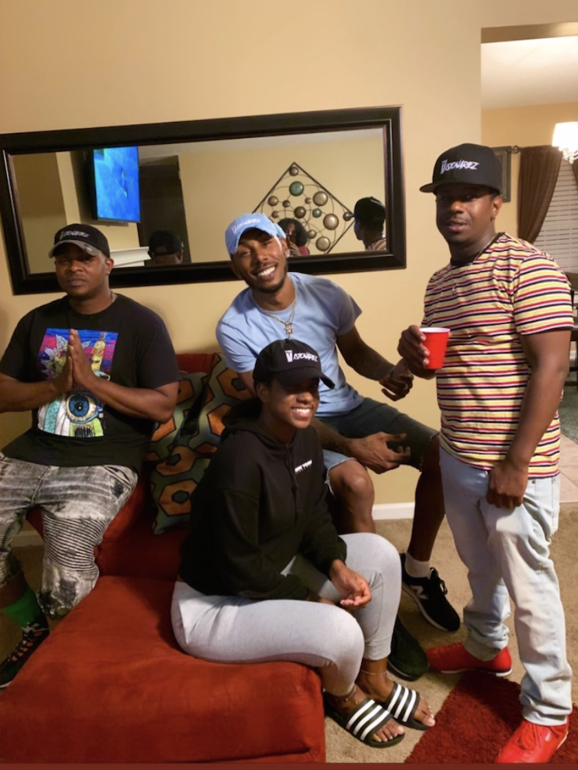 Four young adults enjoying a casual gathering in a living room, with one sitting on a couch, two standing, and one kneeling on the couch, smiling and holding a red solo cup, wearing casual clothes and caps, with a mirror and wall decor in the backgro