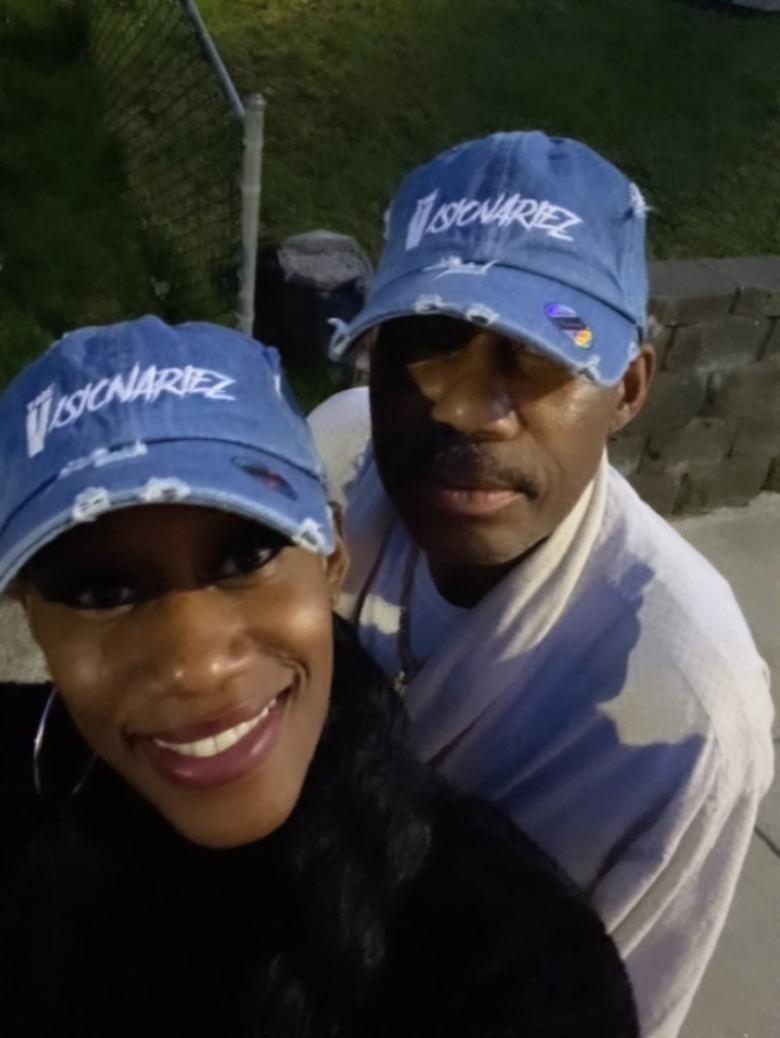 A woman and a man taking a selfie outdoors at night, both wearing blue baseball caps with white text The Visionariez', and the woman smiling at the camera.