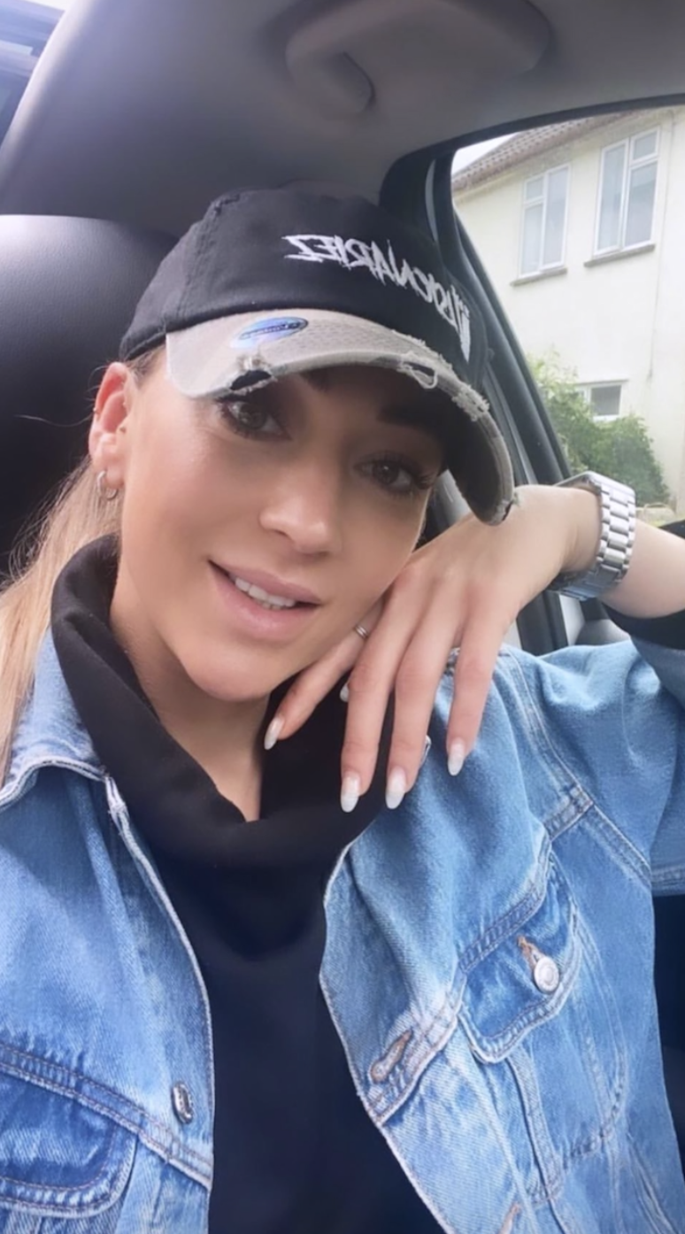 A young woman with light skin, long hair, and makeup, wearing a black cap with distressed details and white lettering, a black hoodie, and a blue denim jacket. She is sitting in a car, smiling and resting her hand on her face, with a house visible ou