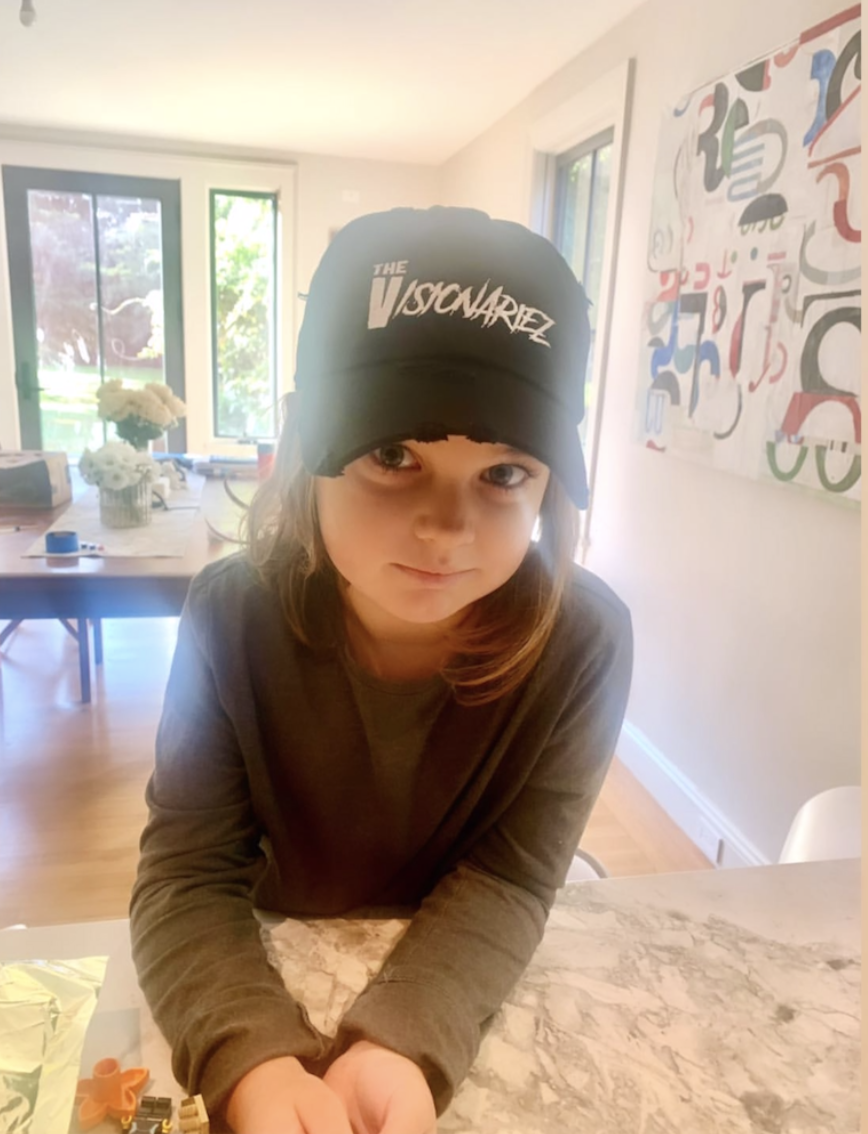 A young girl with brown hair wearing a black baseball cap that says 'The Visionariez' in white text, standing inside a well-lit home with a dining table and abstract art on the wall in the background.