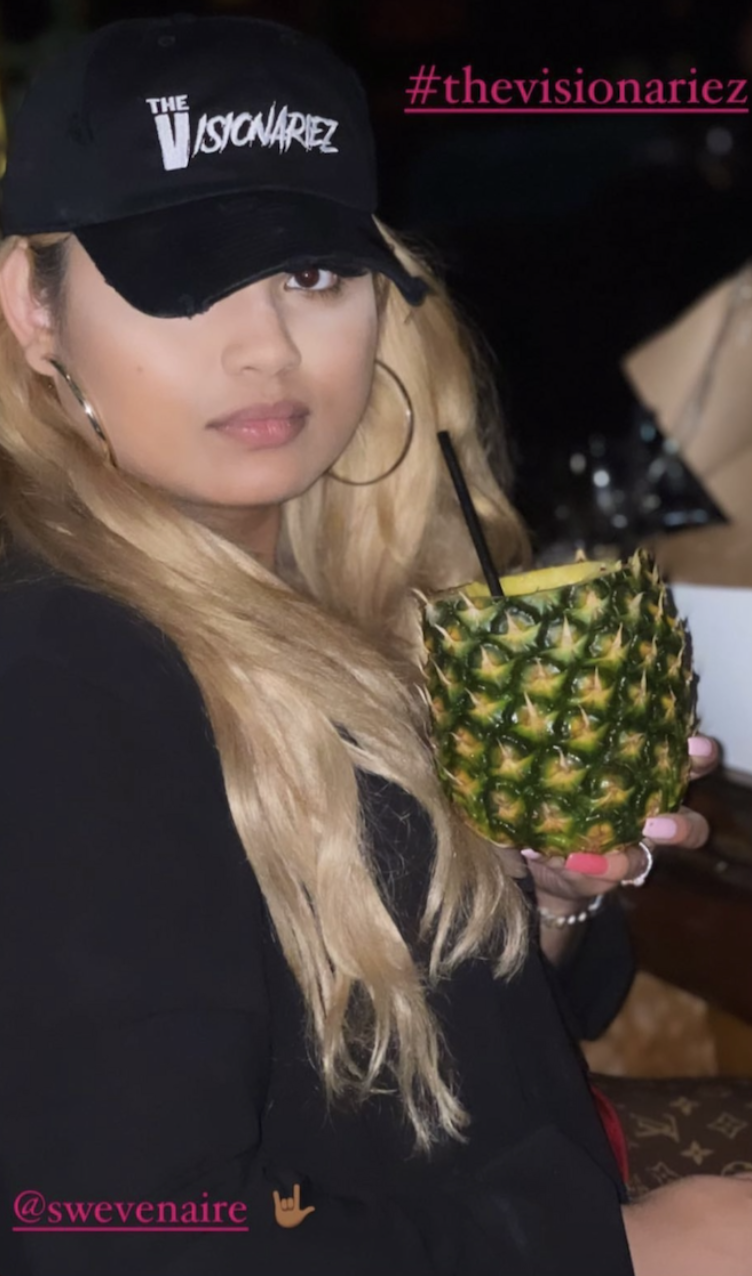 A woman with long blonde hair wearing a black cap with white logo and large hoop earrings holding a hollowed-out pineapple with a drink inside and a straw. She is dressed in black and has pink nail polish. The Visionariez