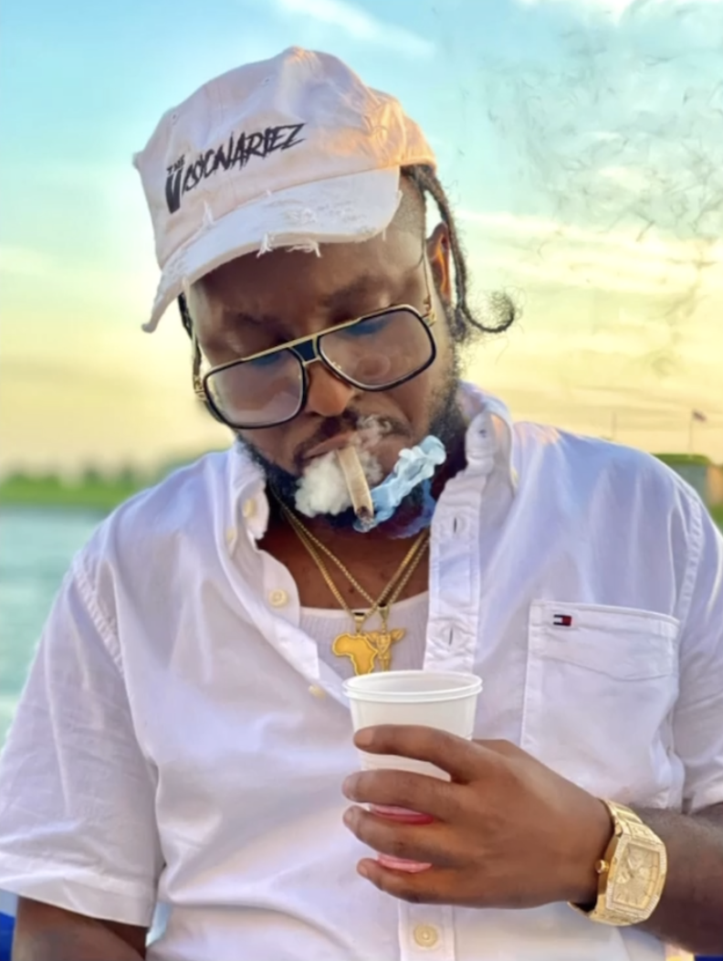 A man with dreadlocks wearing a white cap, glasses, gold jewelry, and a white shirt, holding a cup and smoking. The Visionariez