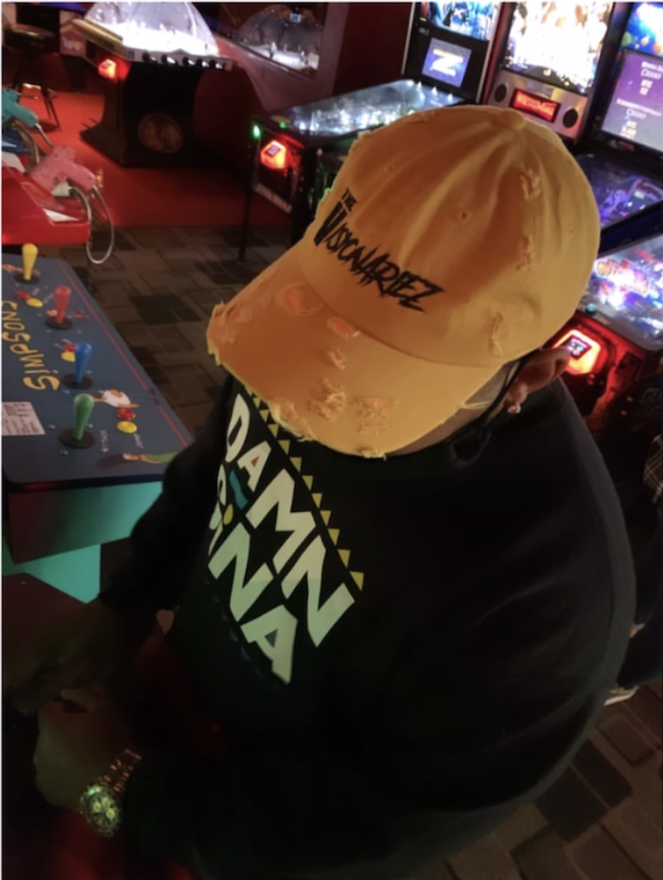 Person wearing a beige distressed cap with black text that reads 'The Visionariez', sitting in an arcade room with pinball machines and arcade games in the background.