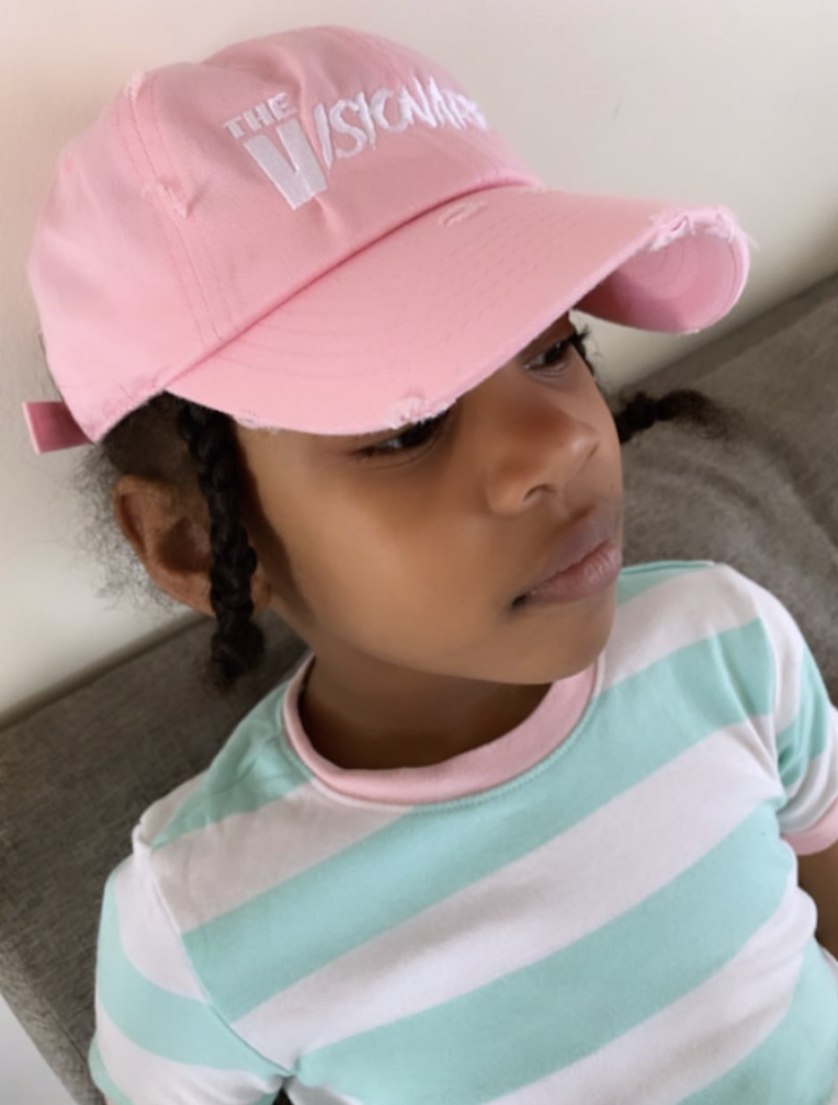A young girl with braided hair wearing a pink cap and a striped shirt sitting on a couch.
