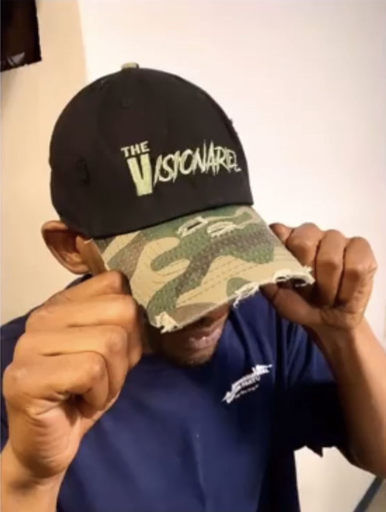 Person wearing a black baseball cap with the phrase 'The Visionariez' and a camouflage brim, pulling the brim down with both hands.