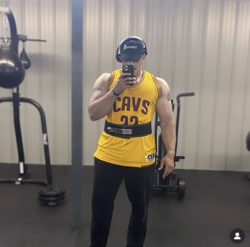 Person taking a mirror selfie in a gym. They are wearing a Cleveland Cavaliers basketball jersey, black pants, and a weighted belt. They have a hat and headphones on. The Visionariez