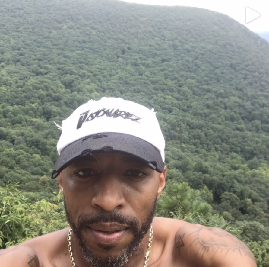 A man with a beard and tattoos on his shoulders taking a selfie outdoors in front of lush green hills or mountains. The Visionariez