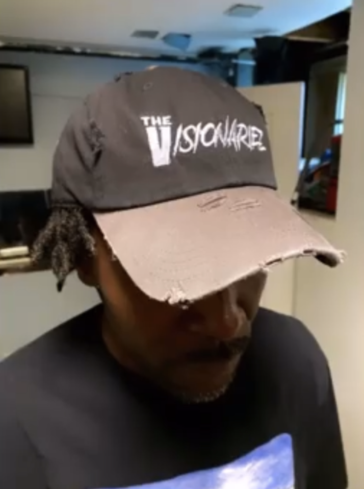 Person wearing a weathered black and pink cap with white text that reads "The Visionariez'" and a black T-shirt, indoors with a blurry background.