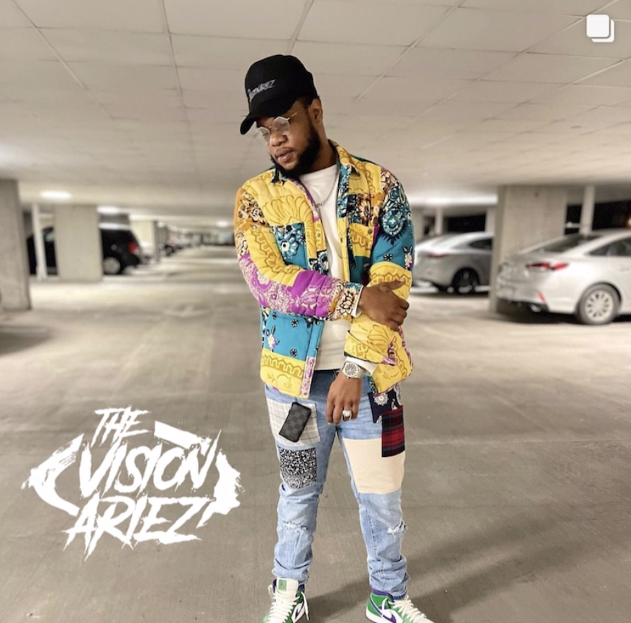 A young man posing in a parking garage, wearing a colorful patterned jacket, distressed jeans with patches, a black cap, sunglasses, and sneakers, with a phone hanging from his pocket and a watch on his wrist. The Visionariez