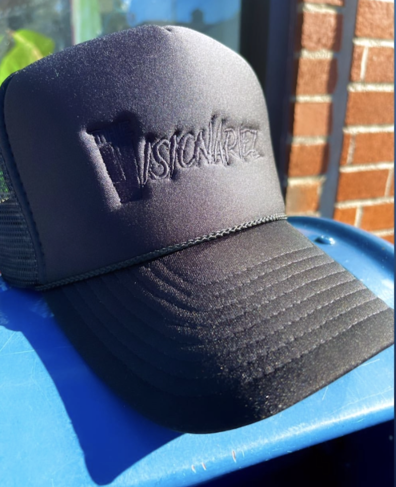 Black trucker hat with embroidered logo reading "The Visionariez" on the front, resting on a blue surface outdoors with brick wall and greenery in the background.