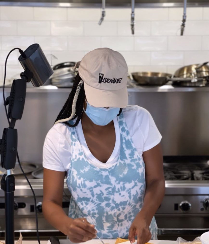 A woman wearing a beige baseball cap with the text 'The Visionariez', a face mask, and a blue and white tie-dye apron in a professional kitchen, preparing food with stainless steel cookware in the background.