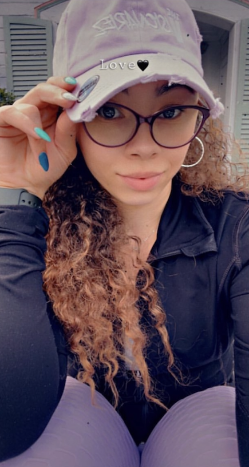 A young woman with curly hair, wearing glasses and a purple cap that says 'The Visionariez'. She is holding her glasses with one hand and smiling at the camera, dressed in a black jacket and purple pants.