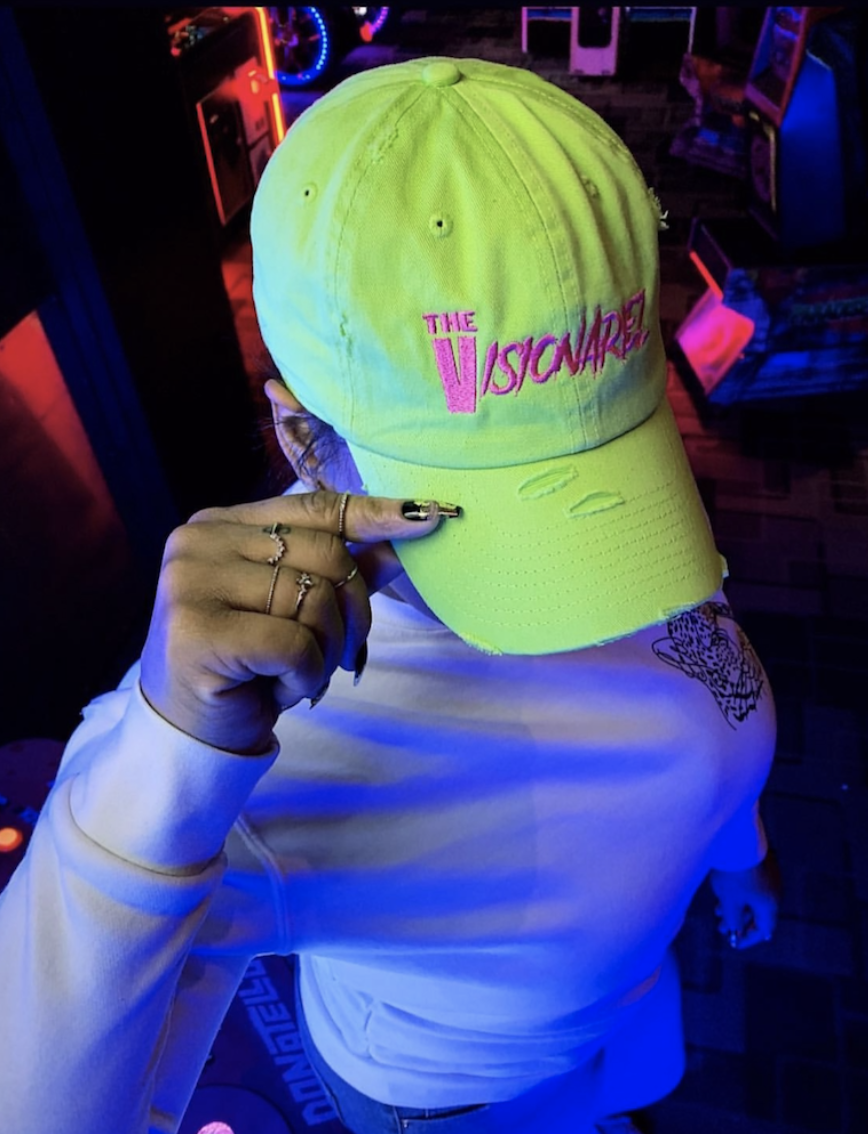Person wearing a bright yellow-green baseball cap with pink embroidered text reading 'The Visionariez' holding the bill of the cap with their index finger, in a neon-lit arcade setting.