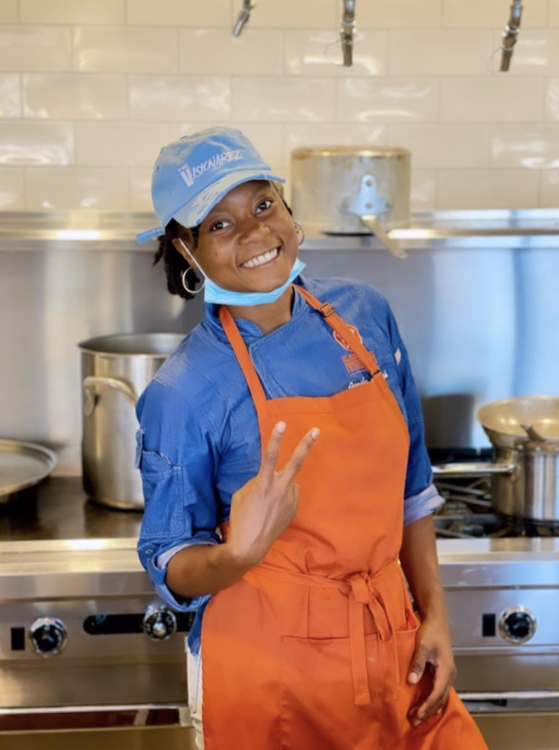 A young woman in a blue chef's uniform, orange apron, and cap smiling and making a peace sign in a commercial kitchen. The Visionariez