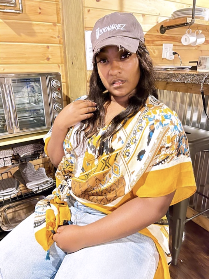 Young woman wearing a gray baseball cap and a yellow-patterned shirt sitting in a wooden-paneled room, with kitchen appliances and utensils in the background. The Visionariez