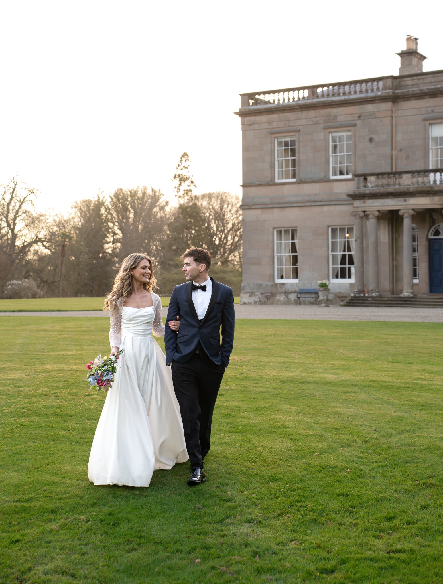 A bride and groom walking arm-in-arm on a grassy lawn near a large historic stone mansion during a wedding photoshoot, with trees in the background and warm sunlight setting.