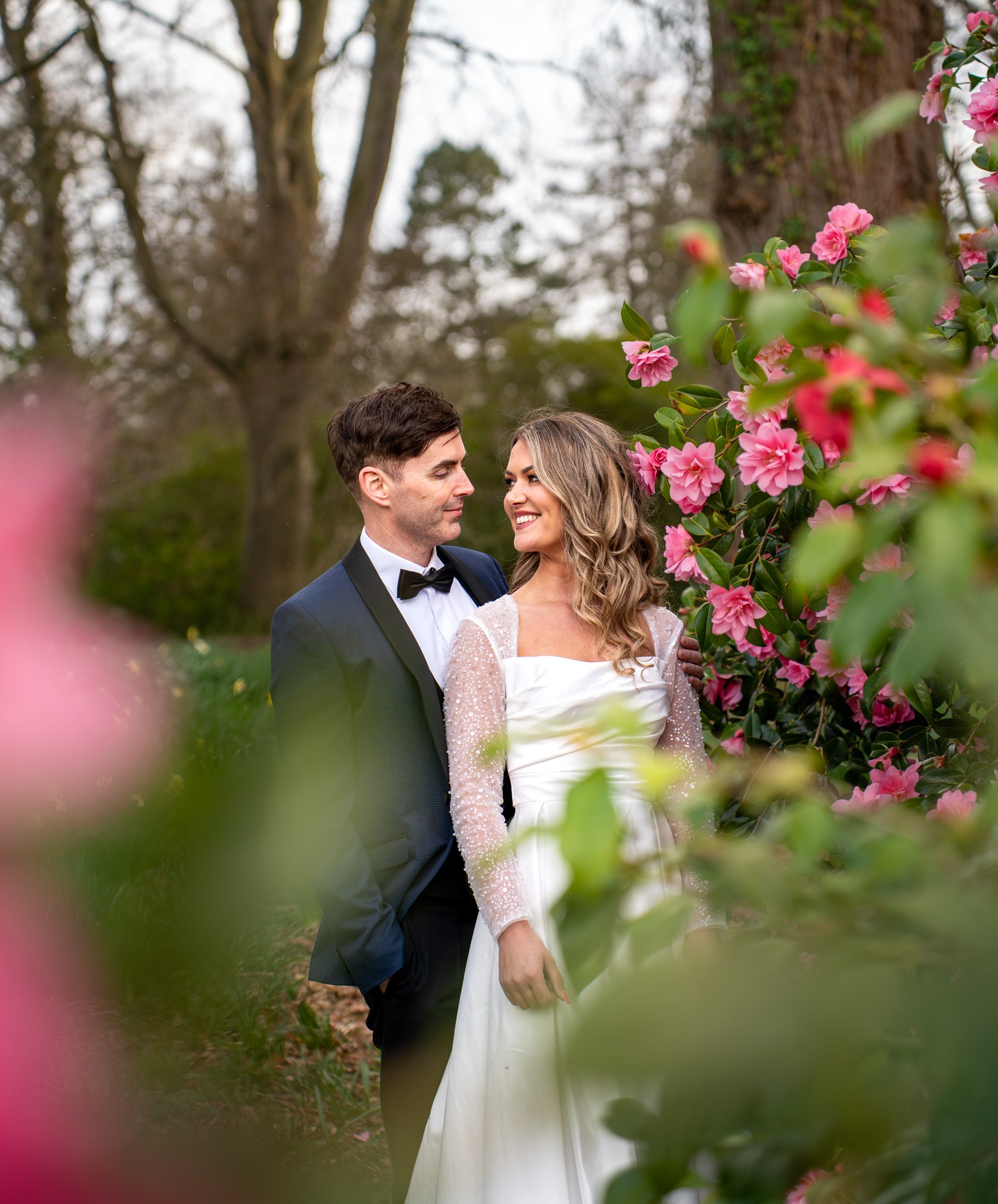 A bride and groom standing close together outdoors surrounded by pink flowering bushes, smiling and looking into each other's eyes.