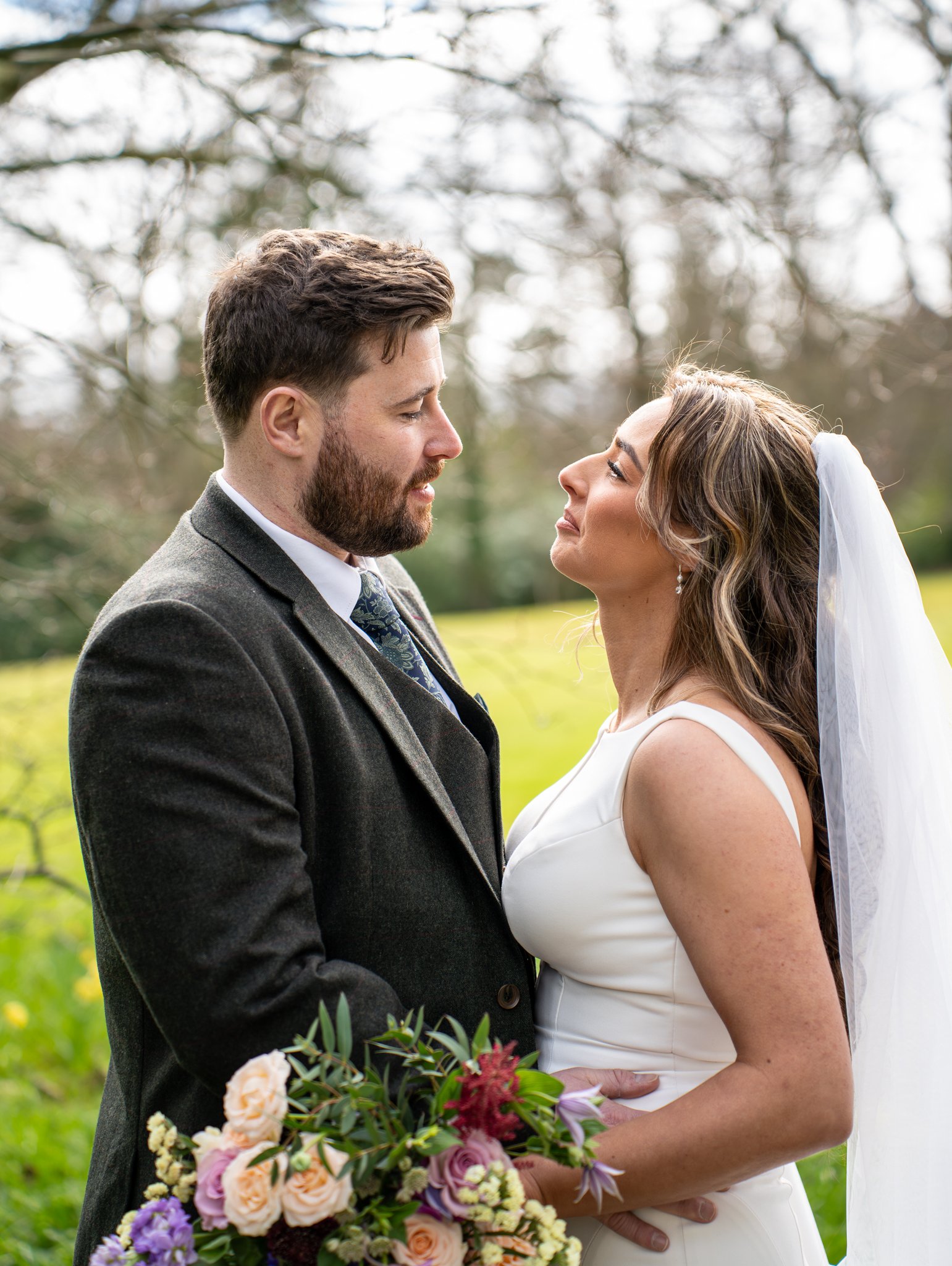 A bride and groom standing outdoors, gazing at each other. The bride is holding a bouquet of flowers, and the groom is dressed in a gray suit with a tie. The setting appears to be a park or garden with trees in the background.