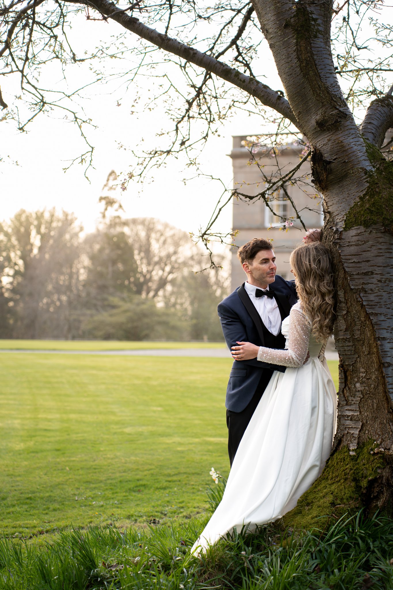 A bride and groom stand close together outdoors near a large tree, with the bride in a white wedding dress and the groom in a tuxedo, looking at each other during sunset with a building in the background.