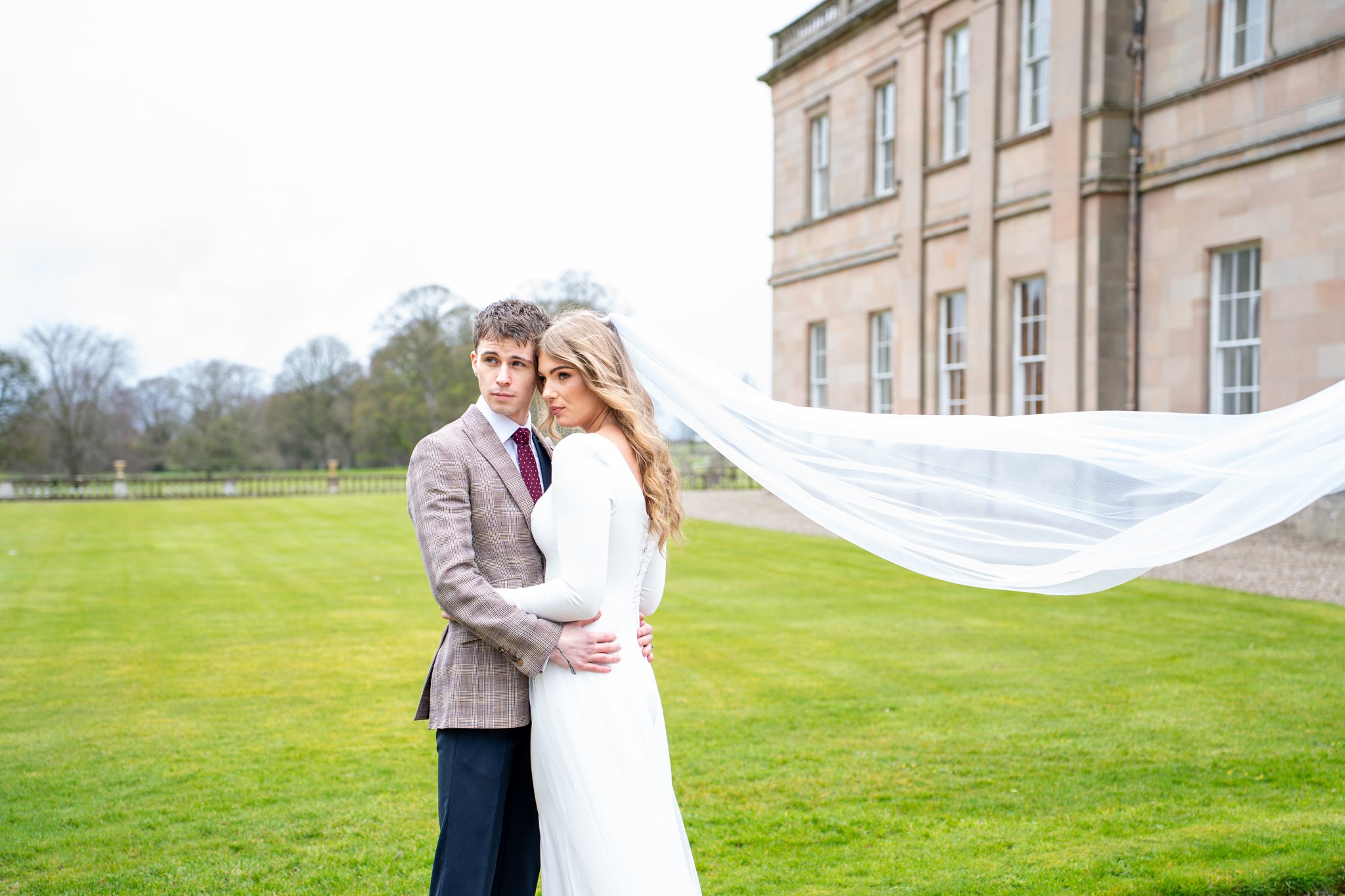 A bride and groom embrace outdoors on a green lawn, with the bride's long veil flowing in the wind, and a historic building in the background.
