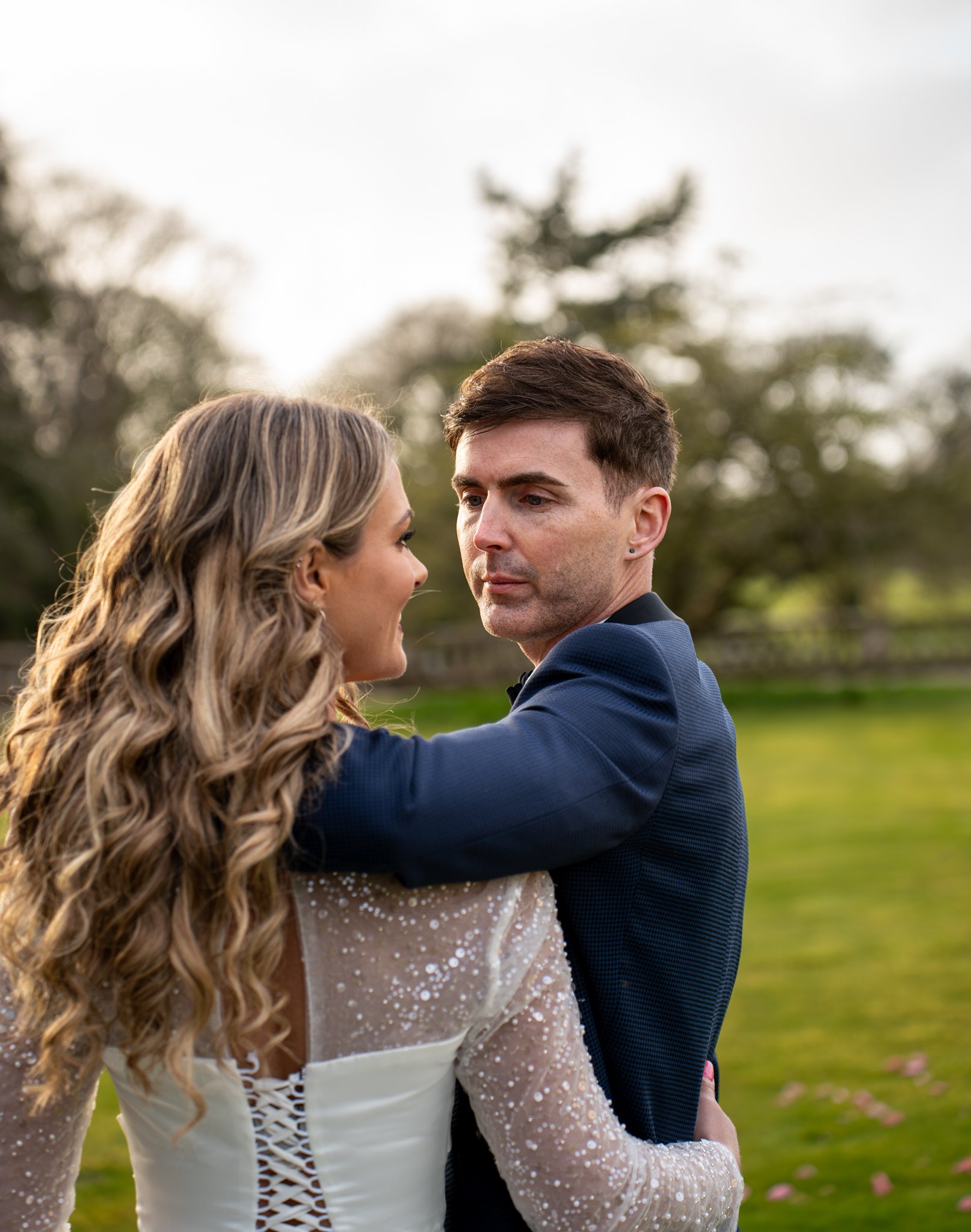 A couple dressed in formal attire embracing outdoors with trees and grass in the background.