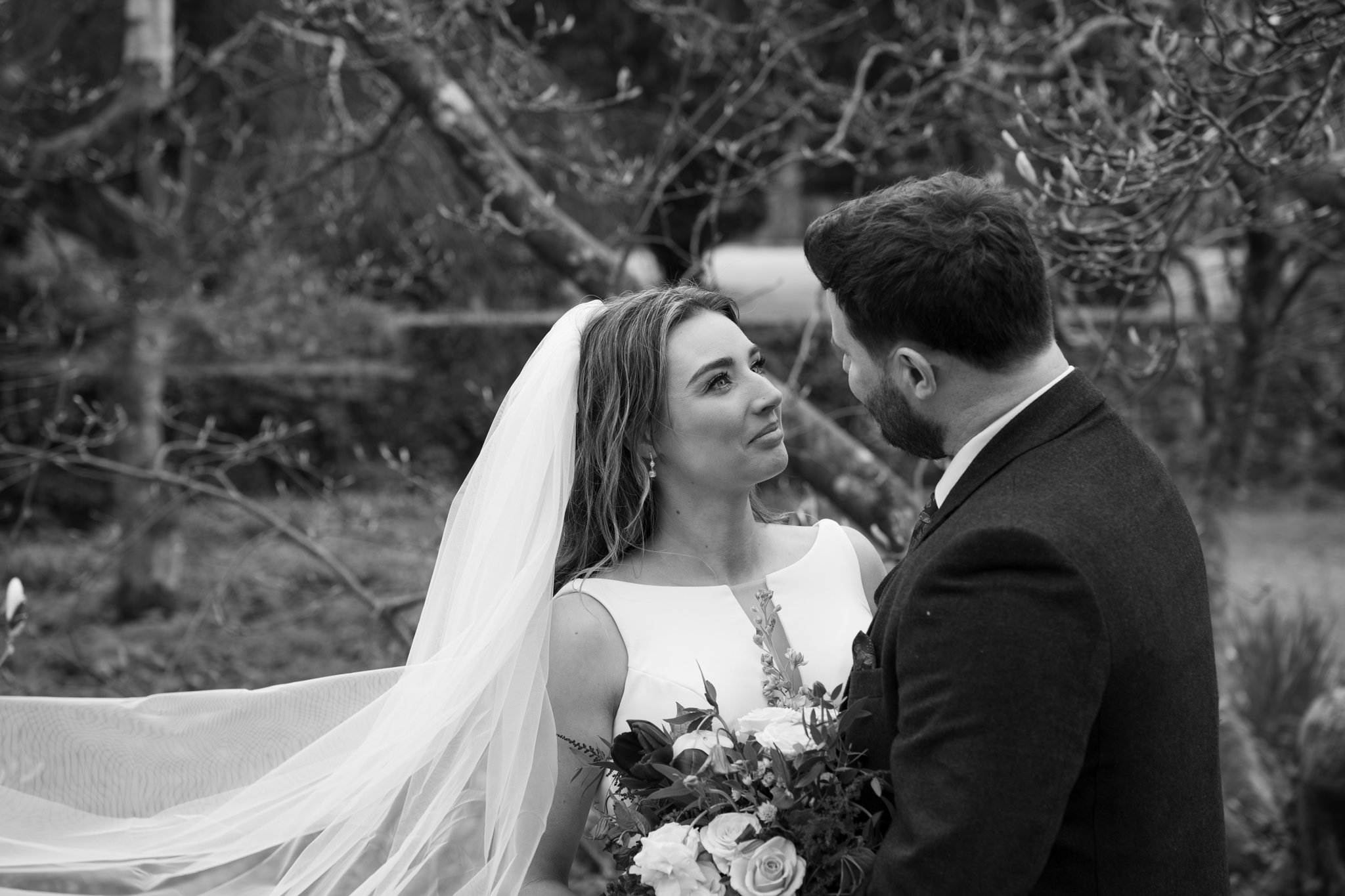 Black and white photo of a bride and groom facing each other outdoors, with trees in the background. The bride wears a wedding gown and veil, holding a bouquet of flowers. The groom wears a suit.