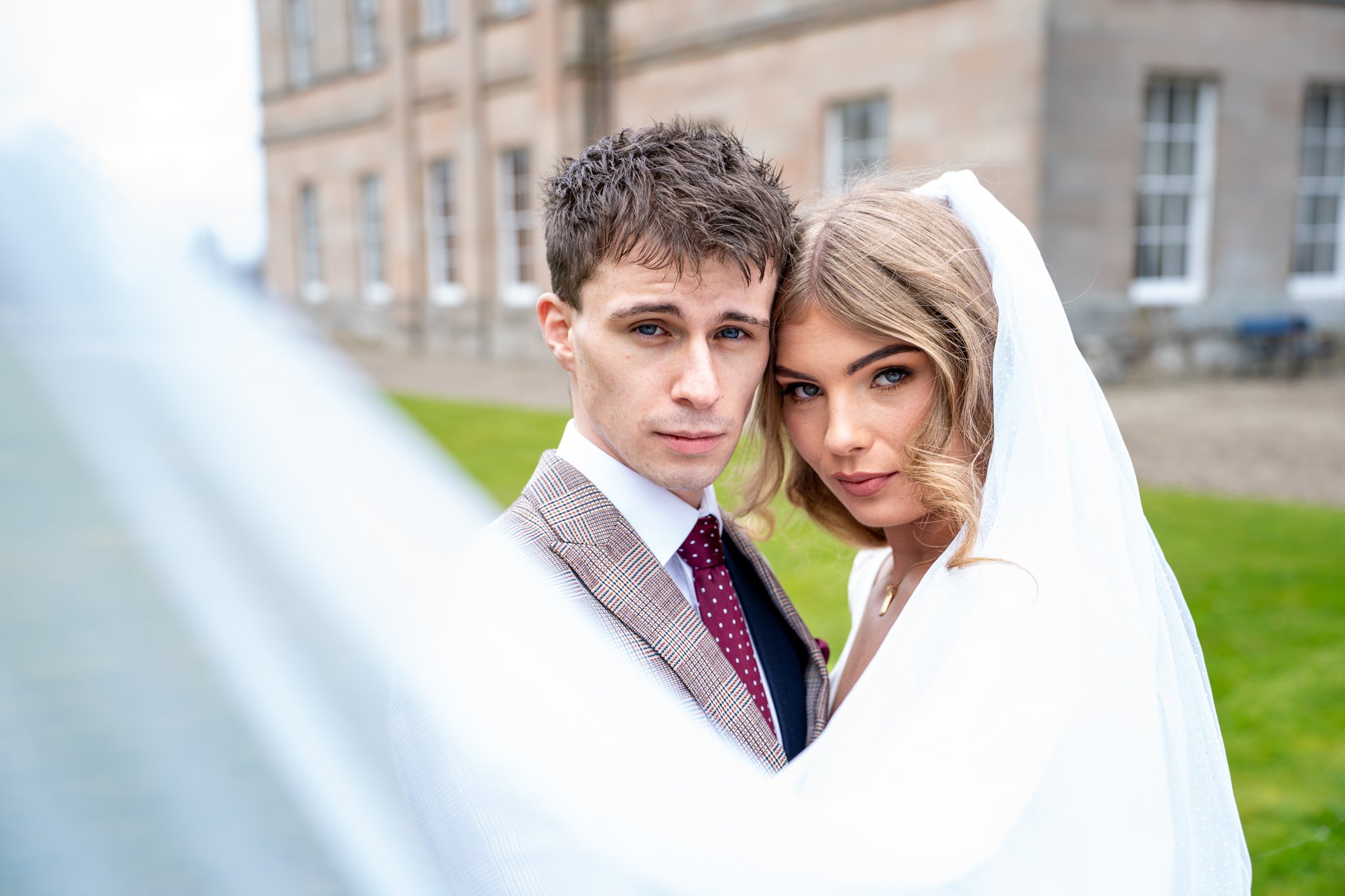 A young bride and groom taking a selfie outdoors, with a blurred historic building and green lawn in the background.