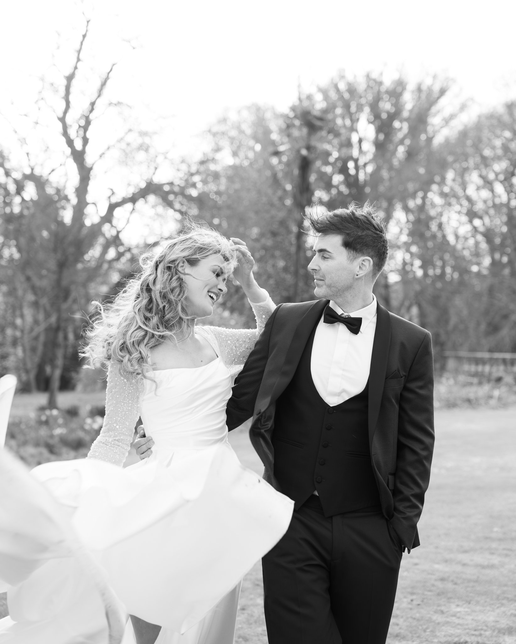 Black and white photo of a newlywed couple outdoors. The bride with curly hair is smiling and holding her dress, while the groom in a tuxedo looks at her with affection. Trees are visible in the background.