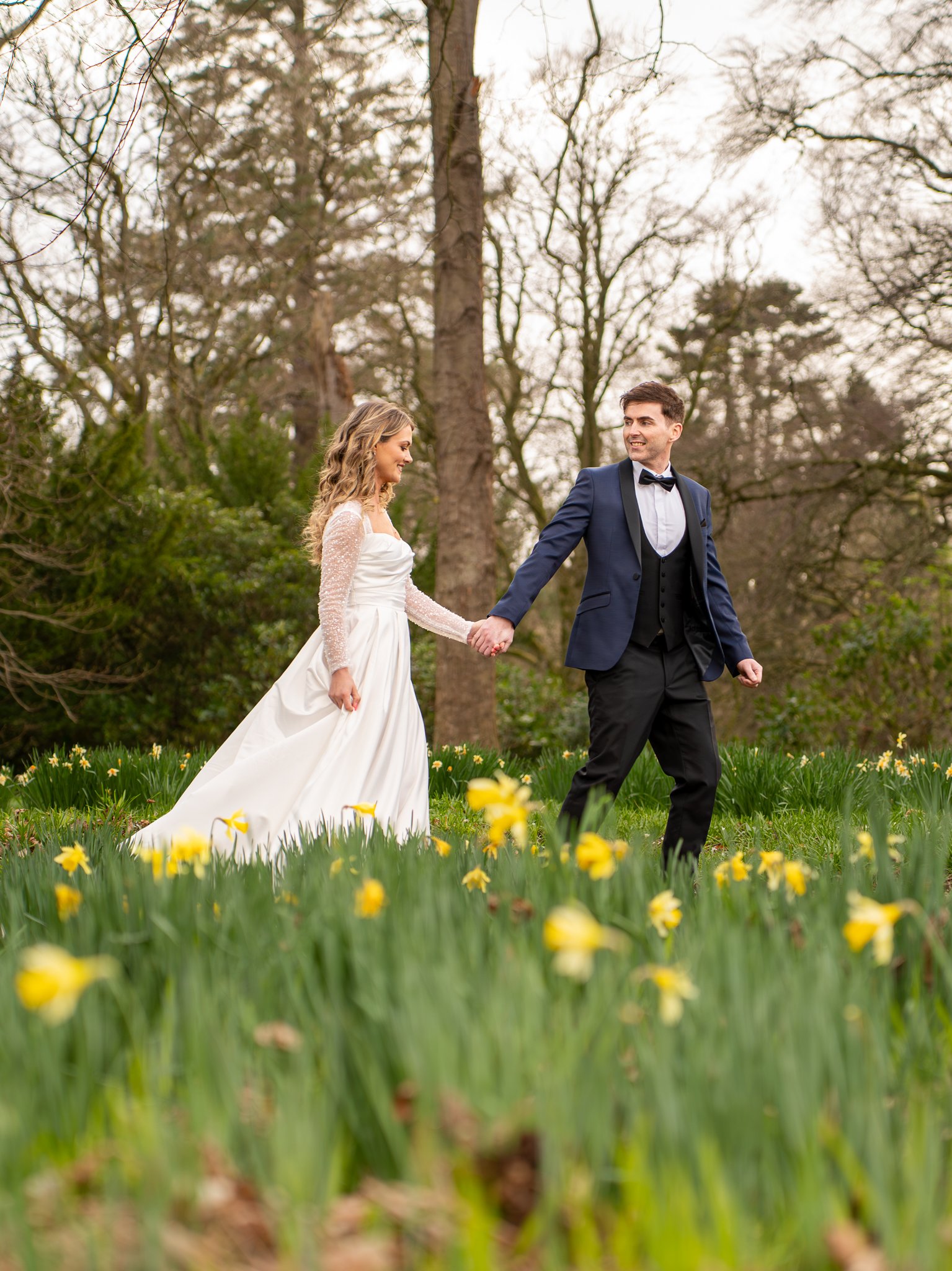 A bride and groom holding hands in a field of yellow flowers during a wedding photoshoot, with tall trees in the background.