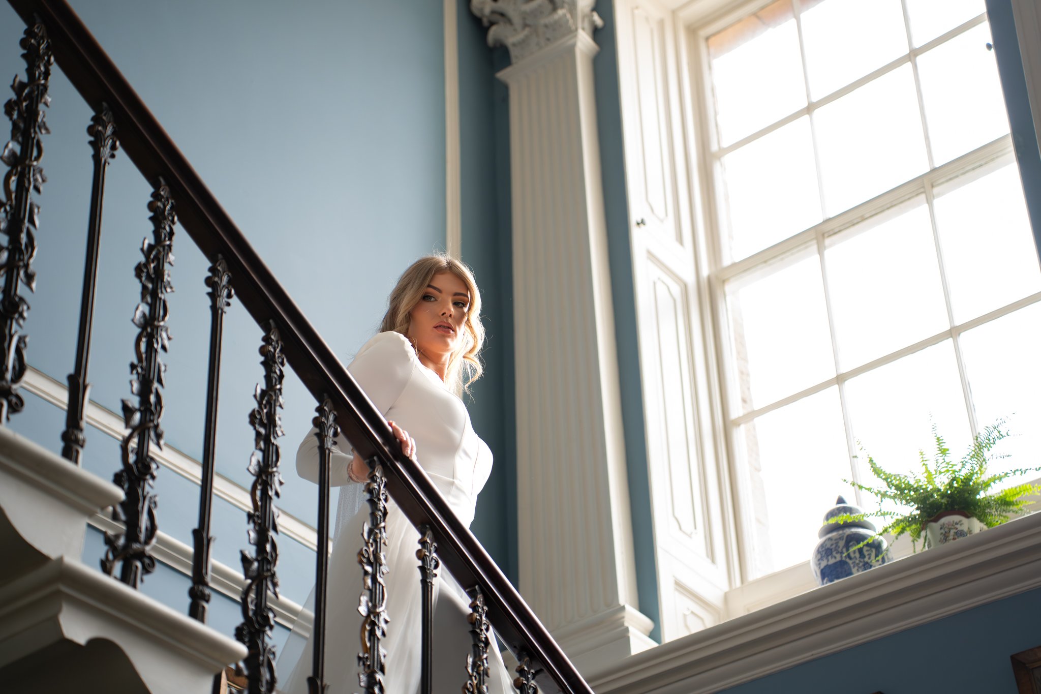 A woman with long blonde hair standing on a staircase, looking over her shoulder at the camera, in a bright room with large window and decorative elements.