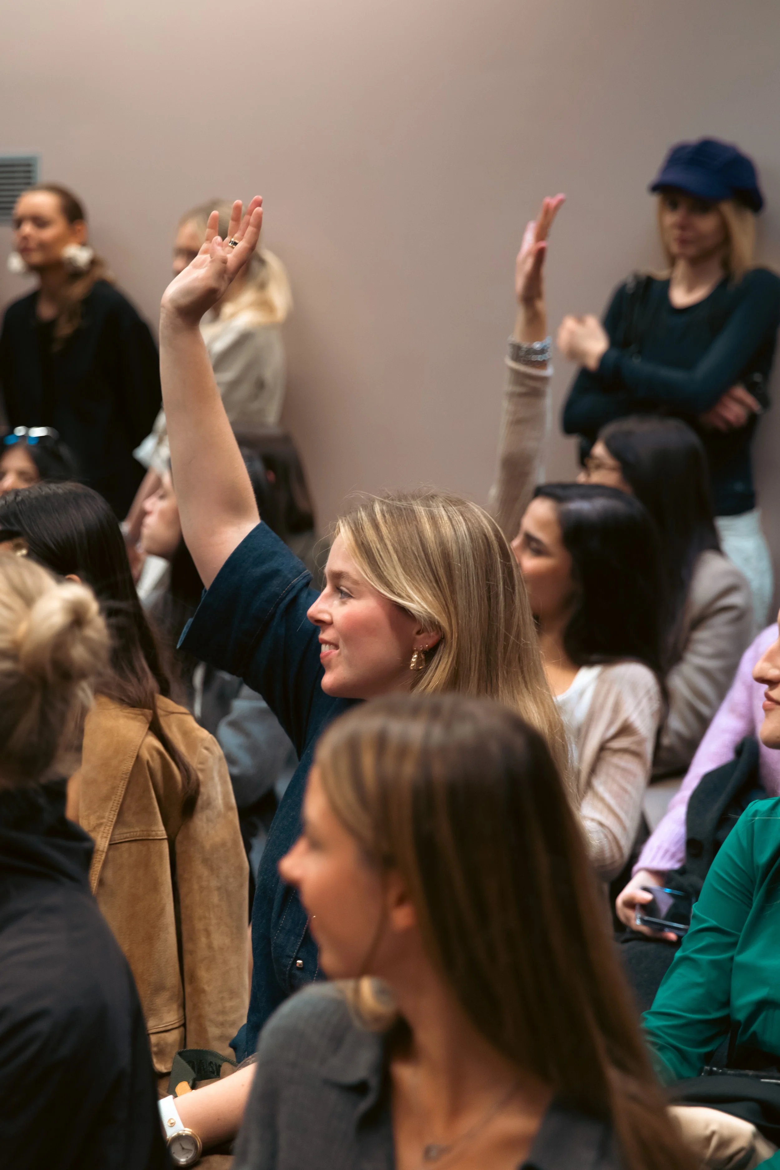 Une femme avec les cheveux blonds, levant la main dans une salle de conférence ou d'événement, entourée d'autres personnes assises ou debout.