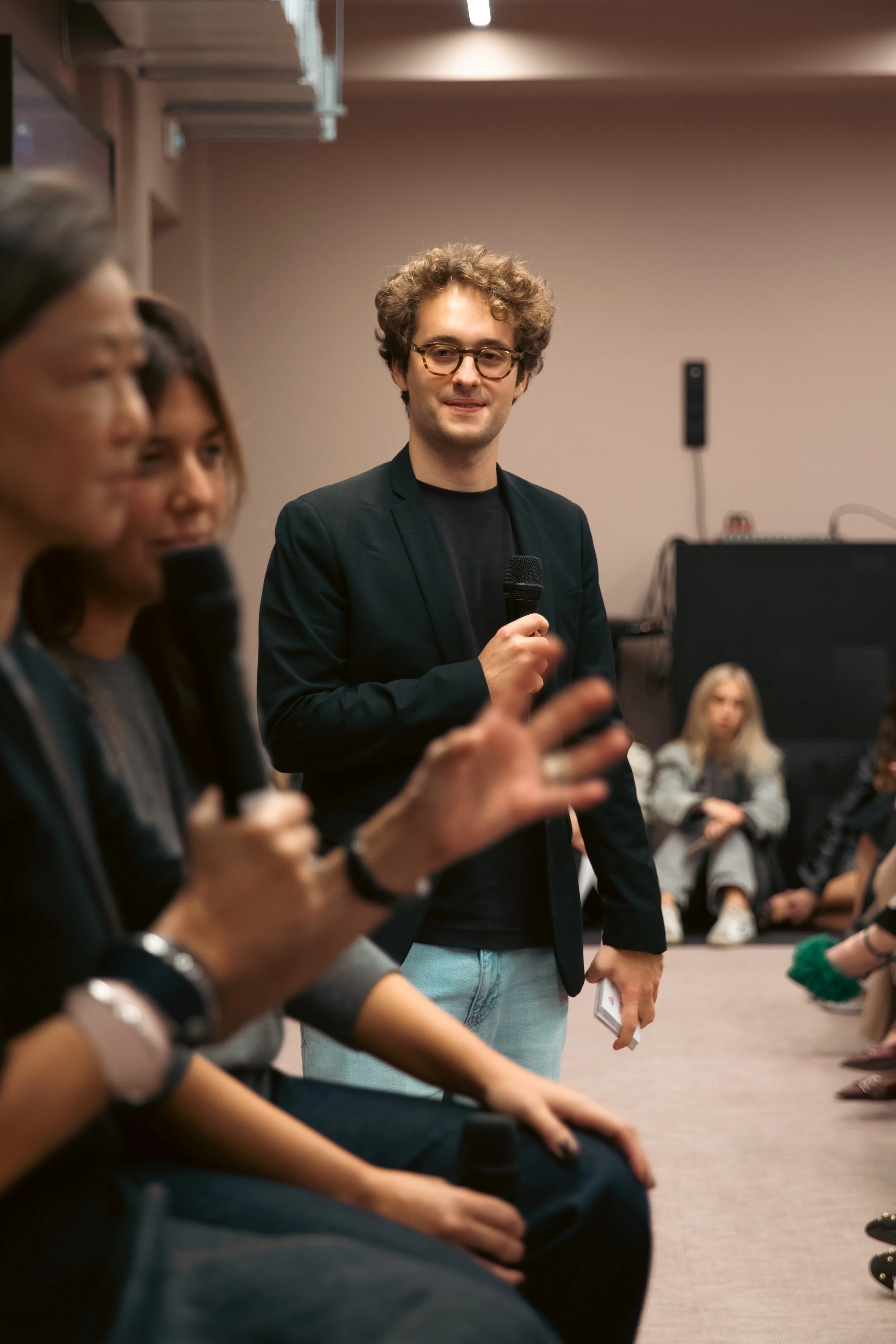 Jeune homme avec des lunettes, portant une veste noire et un t-shirt noir, tenant un micro lors d'une discussion ou d'une présentation, dans une salle en mode conférence ou atelier, avec d'autres personnes assises en cercle.