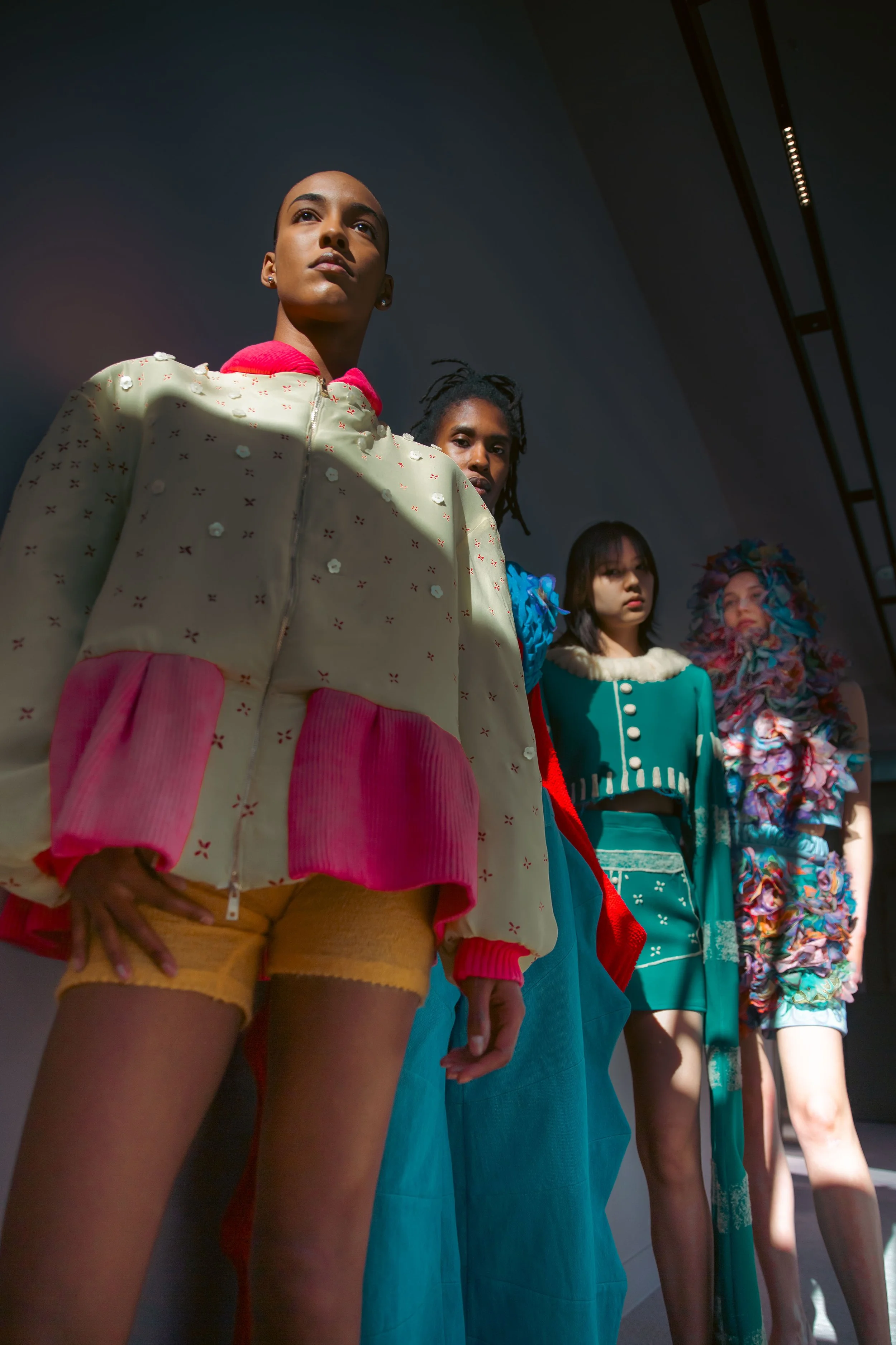 Quatre mannequins féminines portant des vêtements colorés debout, vus de bas en haut, avec un éclairage dramatique.