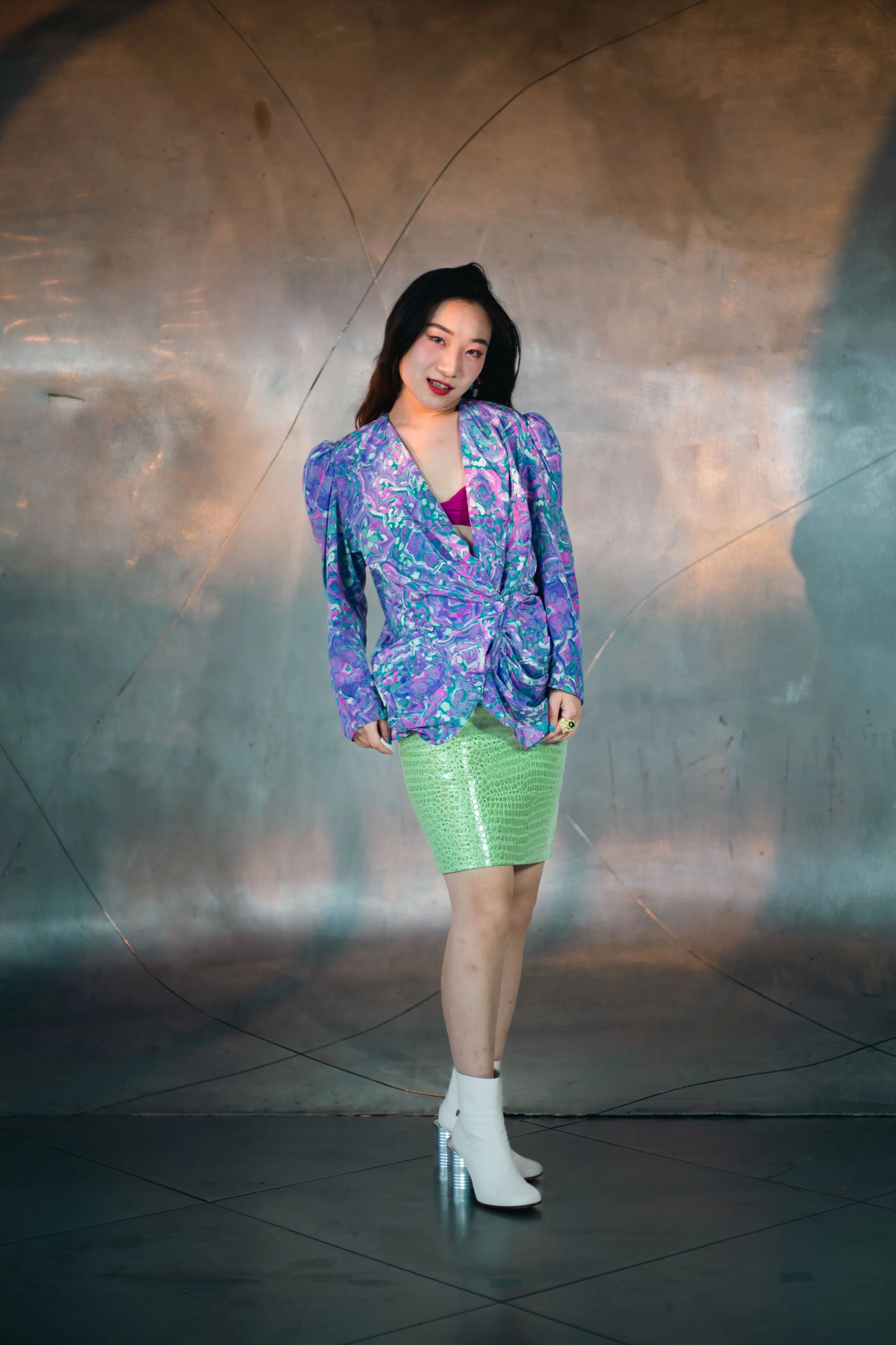 Femme portant un blazer violet avec un motif et une jupe verte en faux crocodile, chaussures bottines blanches à talons transparents, en pose devant un mur métallique