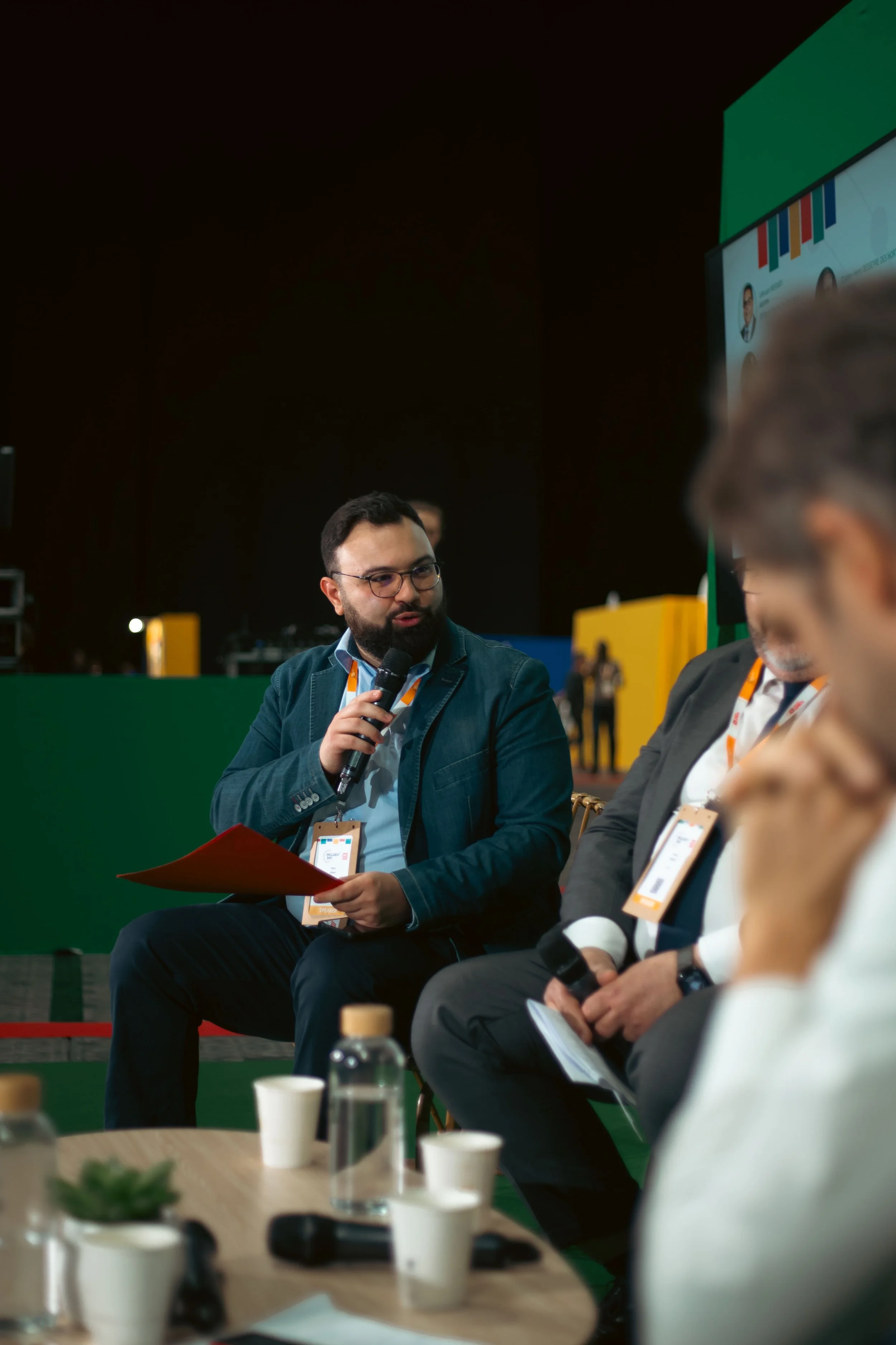 Un homme avec lunettes, barbe et veste en jean, tenant un micro et un dossier rouge, parlant lors d'une conférence ou panel, entouré d'autres personnes assises. En arrière-plan, une scène avec plusieurs écrans et une bannière colorée.