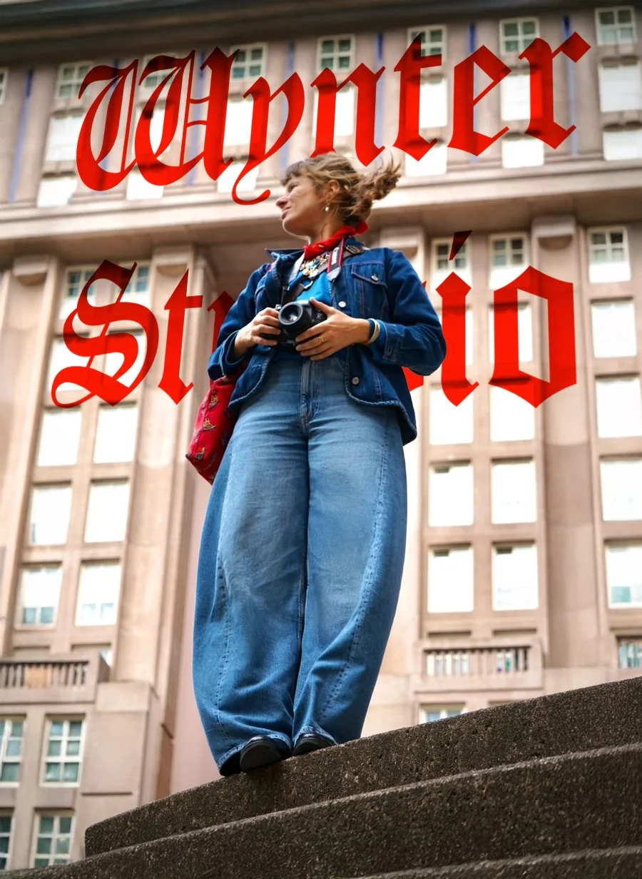 Une jeune femme tenant un appareil photo, portant un jean ample, une veste en jean, avec un bâtiment en pierre en arrière-plan, et le texte en rouge "Vapor Studio" superposé.