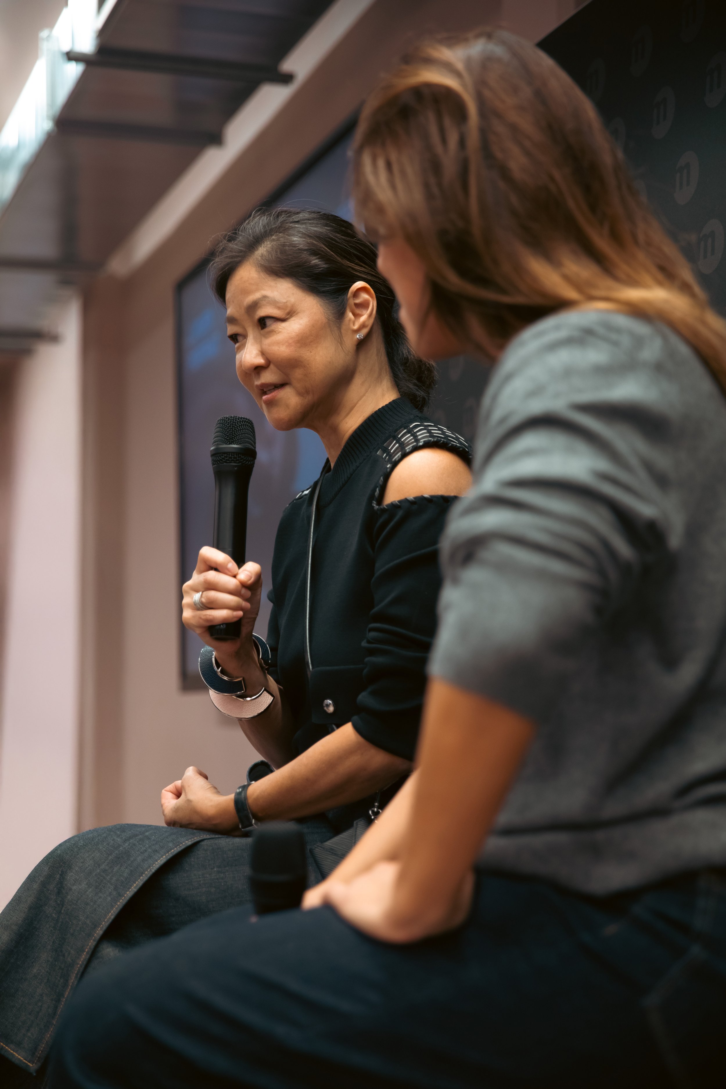 Deux femmes assises en train de parler lors d'une discussion, l'une d'elles tenant un micro, dans une salle intérieure.
