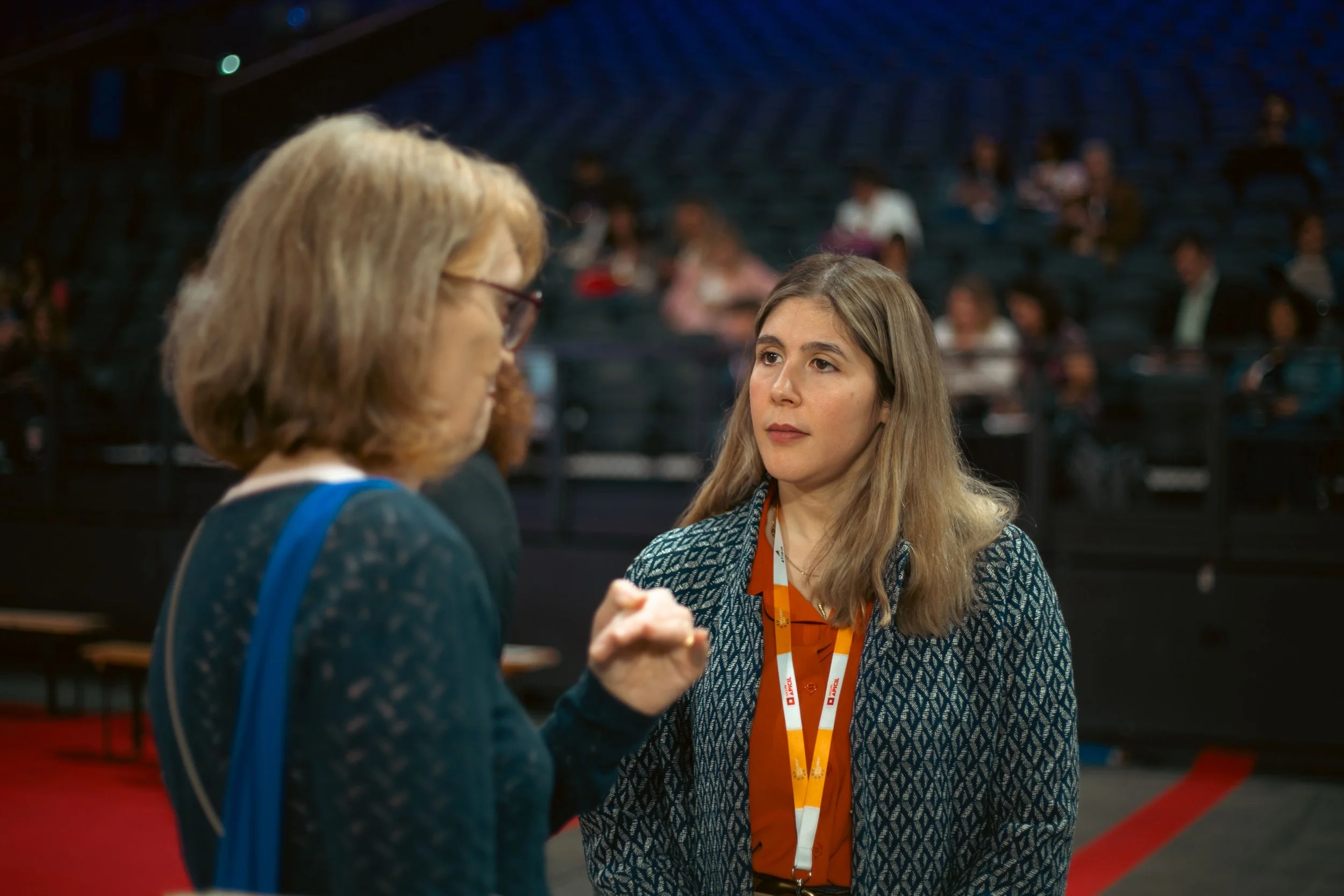 Deux femmes discutent lors d'un événement, une femme avec des cheveux courts et une femme avec des cheveux longs, dans une salle avec plusieurs personnes en arrière-plan.