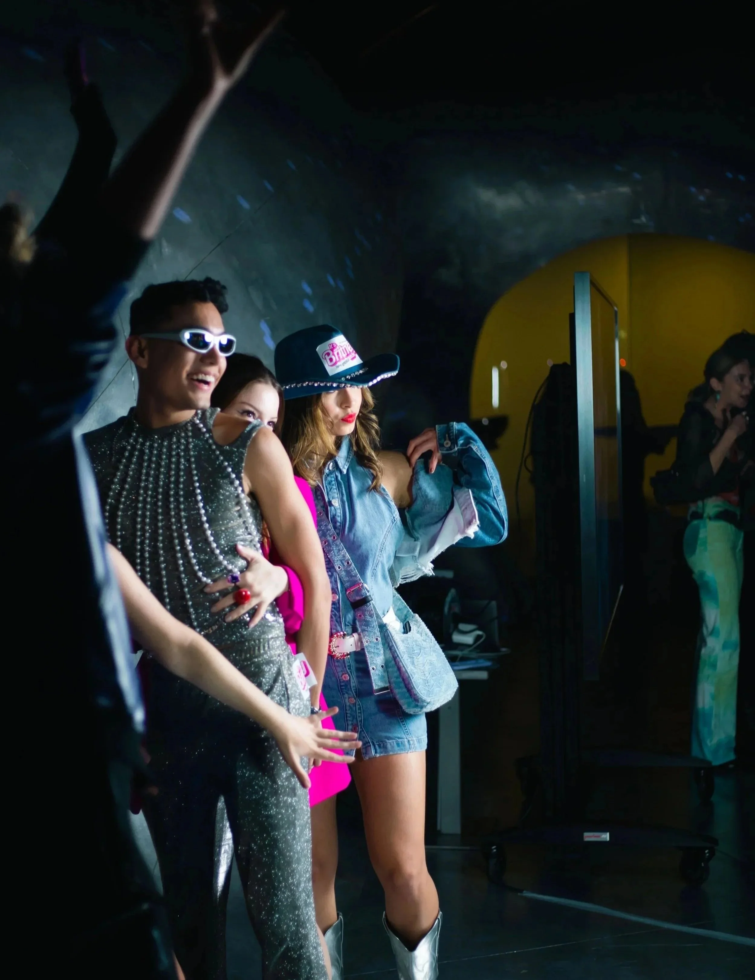 Un groupe de jeunes femmes et un jeune homme habillés de façon colorée et stylée, ils semblent danser ou poser lors d'une fête ou d'un événement dans un lieu sombre avec des lumières colorées.