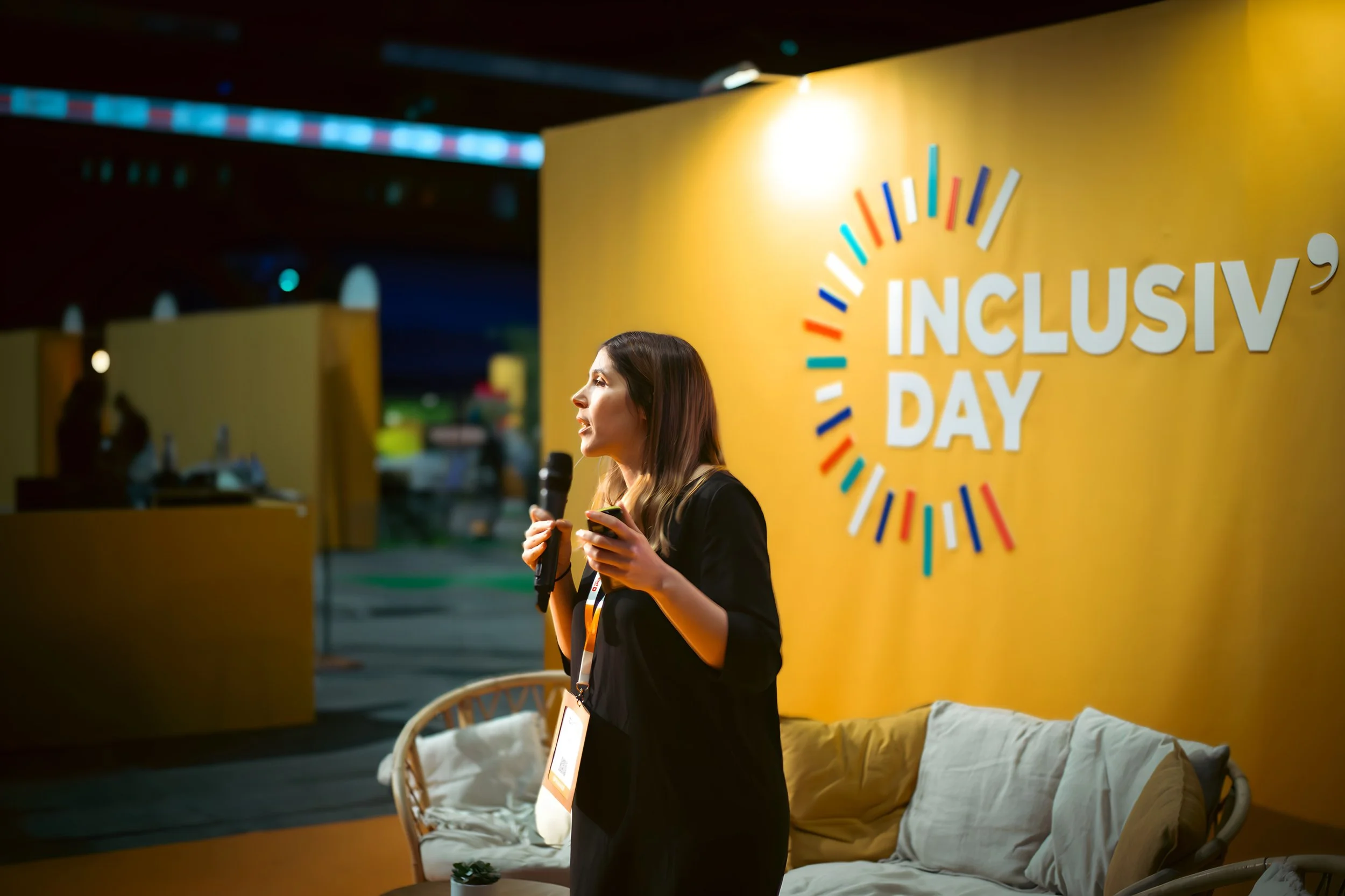 Une femme parle lors d'un événement intitulé 'Inclusiv Day', avec un mur jaune en arrière-plan portant ce nom en gros caractères et un motif de couleur autour.
