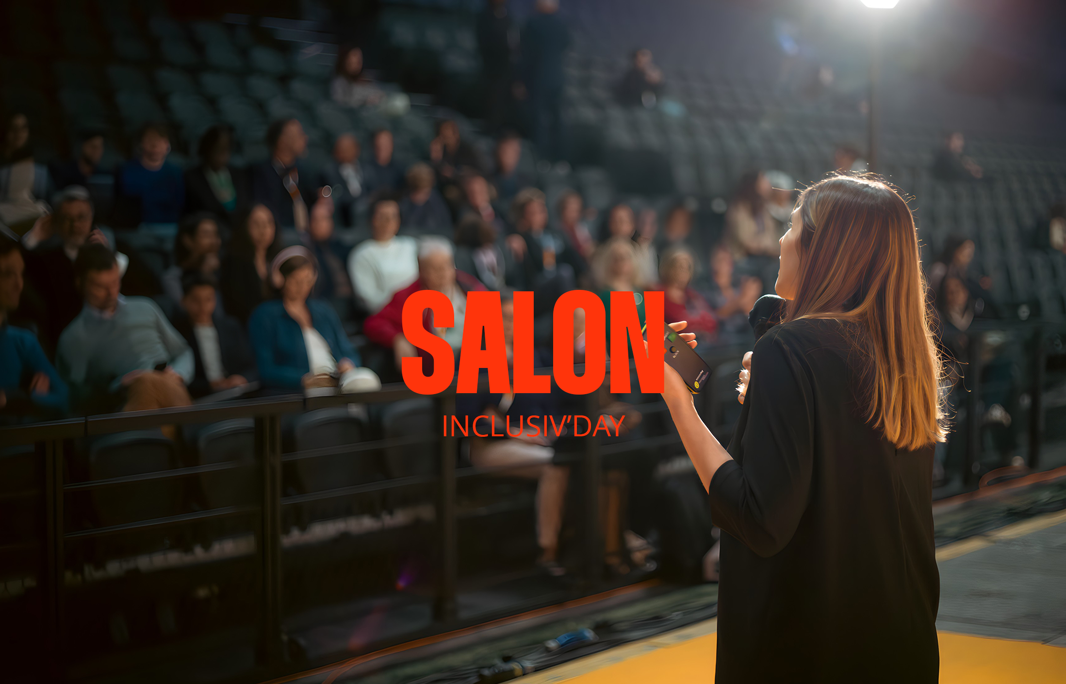 Une conférencière parle lors du salon InclusivDay, avec un public assis en face, sous un éclairage de scène. En avant-plan, une femme avec des cheveux longs, portant un blazer noir, tient un micro et une télécommande.