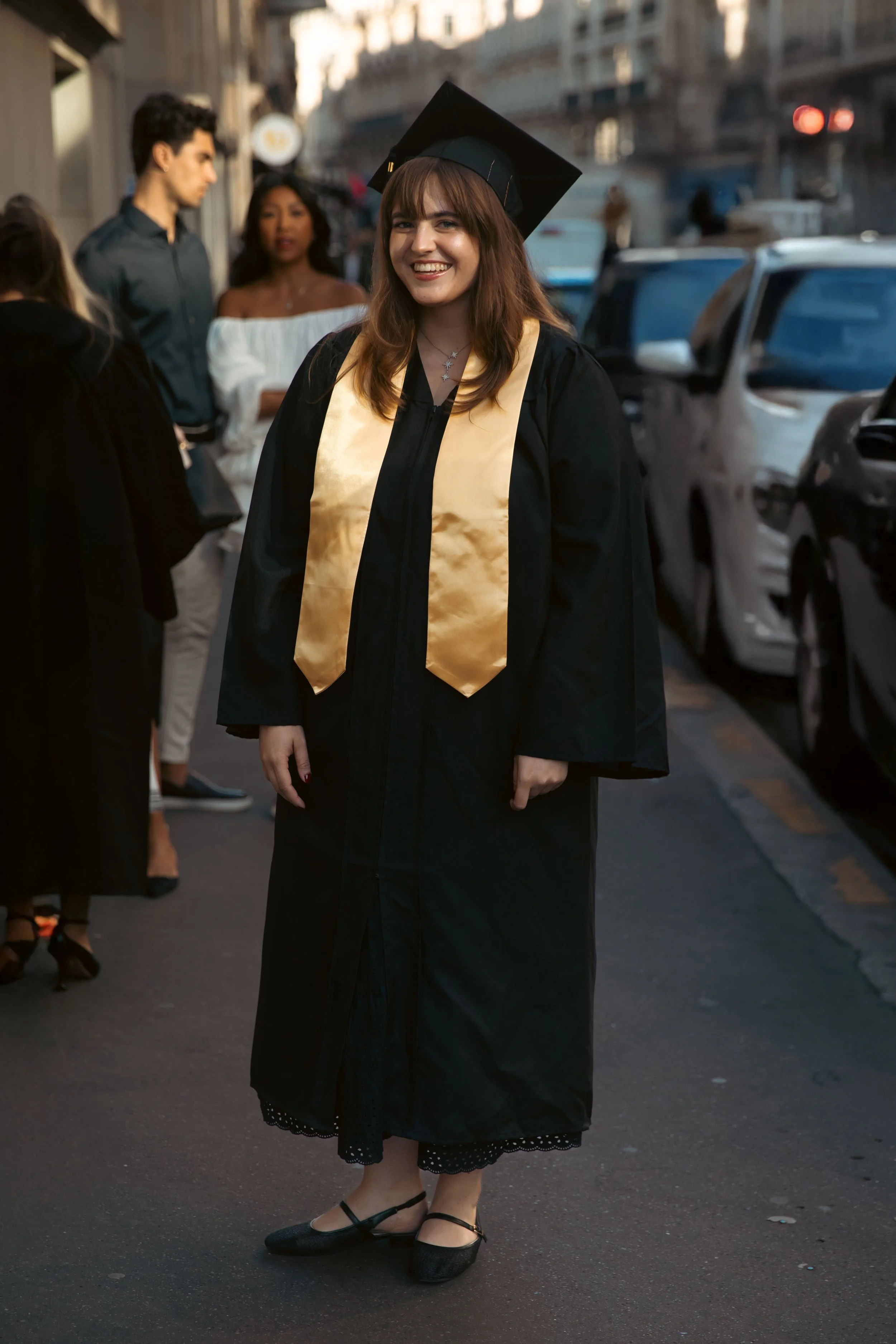 Une jeune femme en robe de diplômée portant une toge noire, un chapeau de cérémonie, une écharpe dorée et des chaussures noires, souriant lors d'une cérémonie de remise de diplôme en plein air.