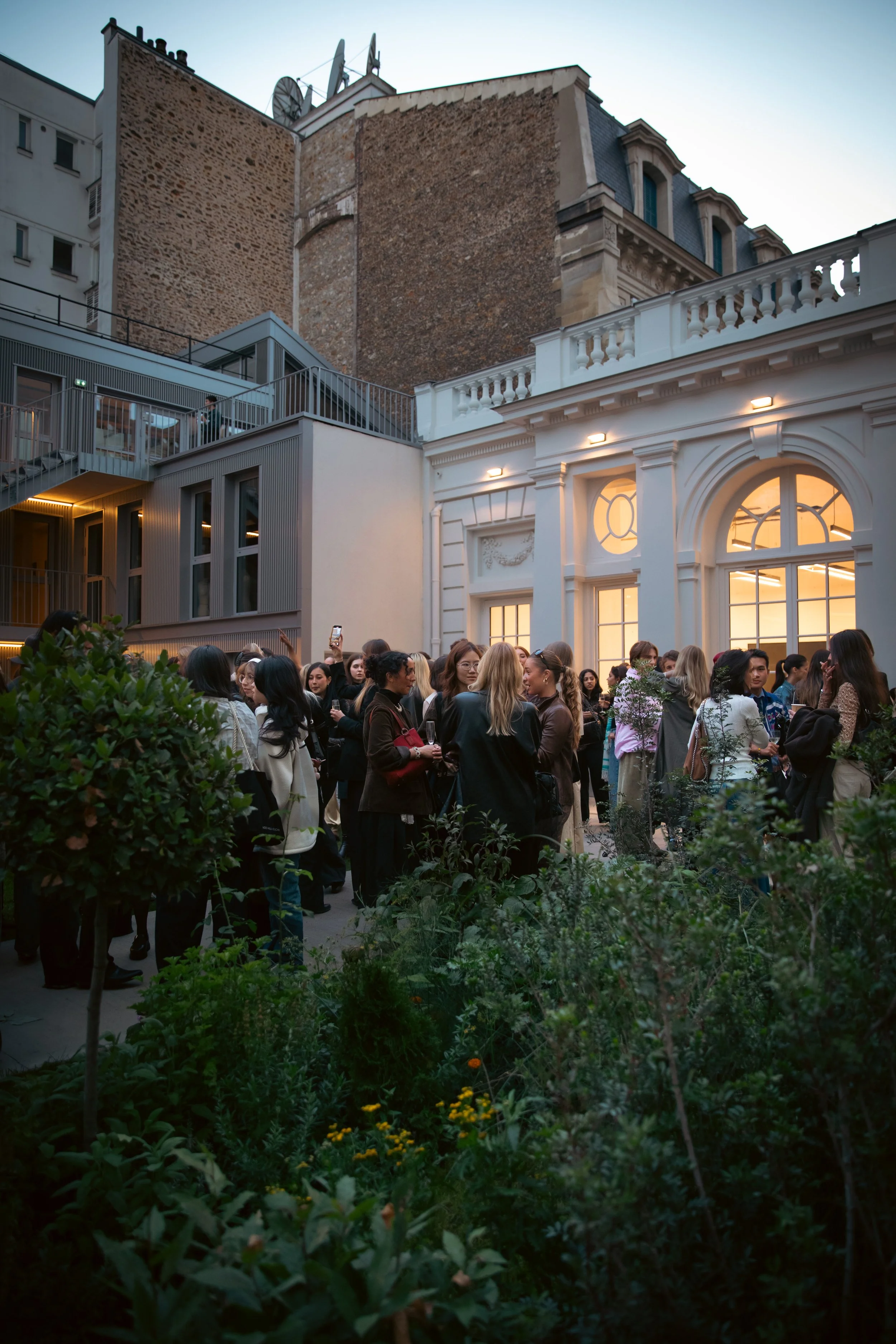 Un groupe de personnes réunies lors d'un événement en intérieur, à l'extérieur d’un bâtiment élégant avec de grandes fenêtres, sous une lumière douce du crépuscule, entouré de végétation