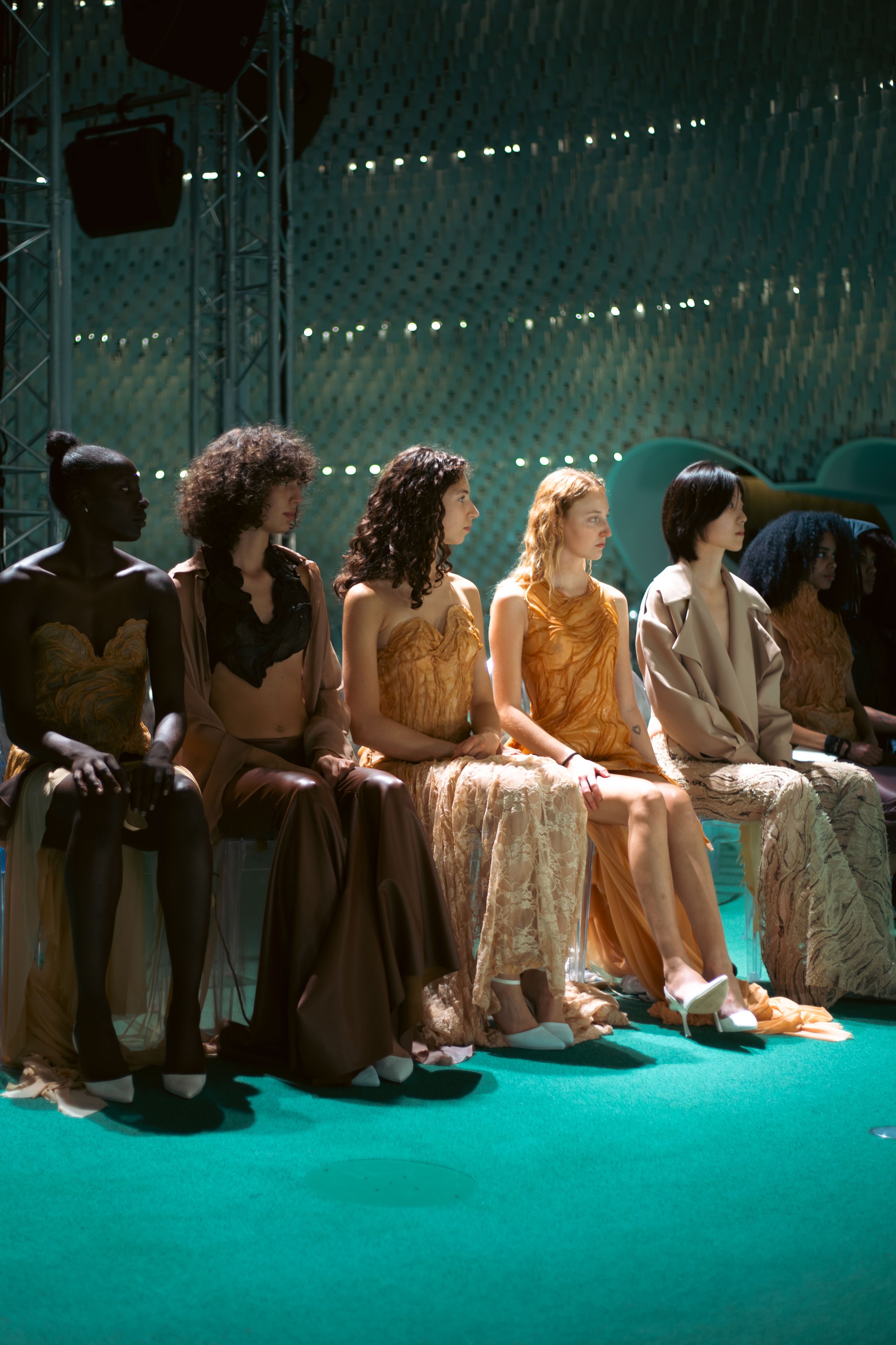 Six femmes assises sur des chaises lors d'un événement de mode, portant des tenues élégantes et variées, dans un environnement intérieur avec éclairage dramatique.